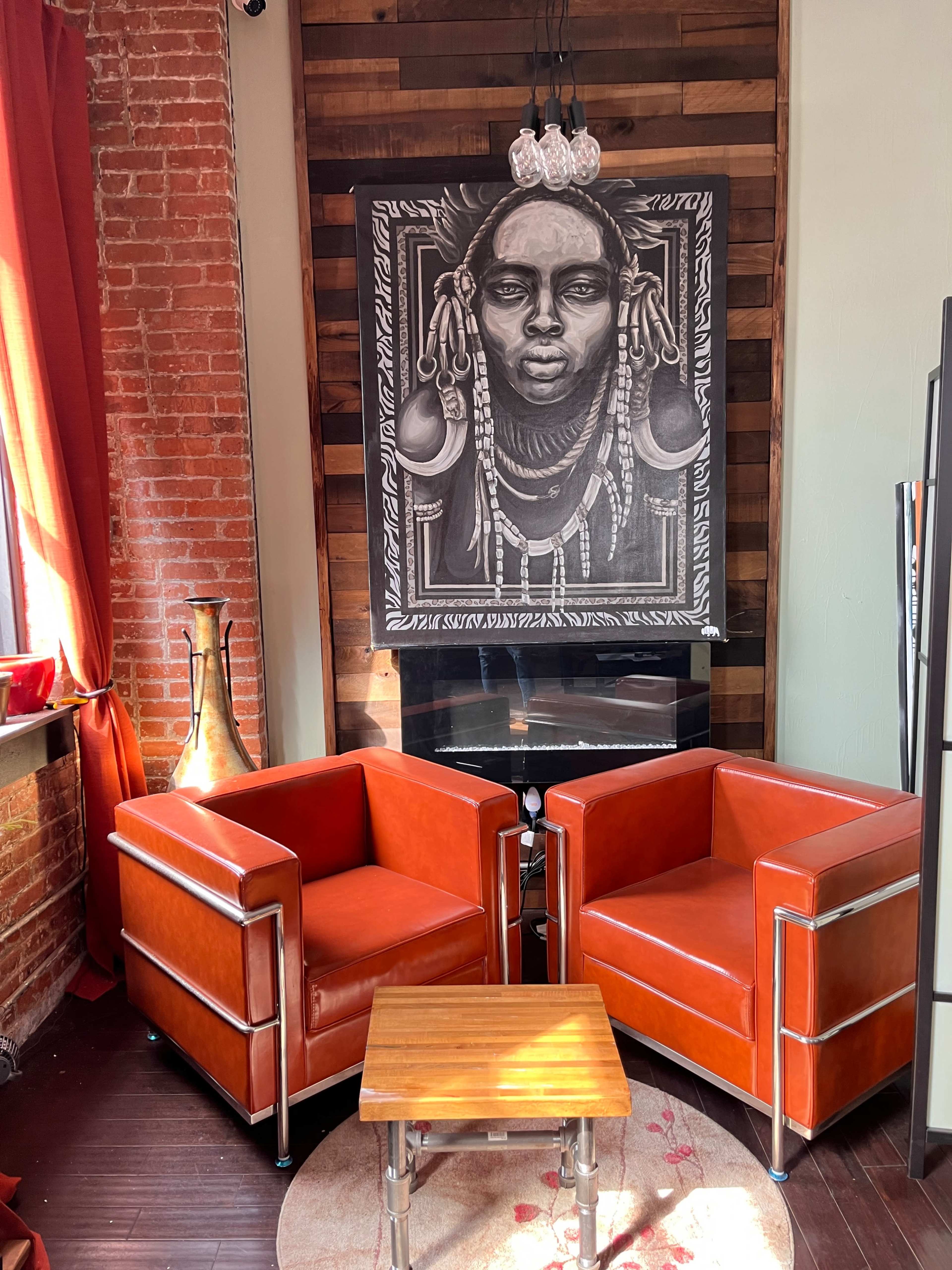 Two orange chairs facing each other with a small wooden table between them, set against a backdrop of a black and white artwork and a brick wall.