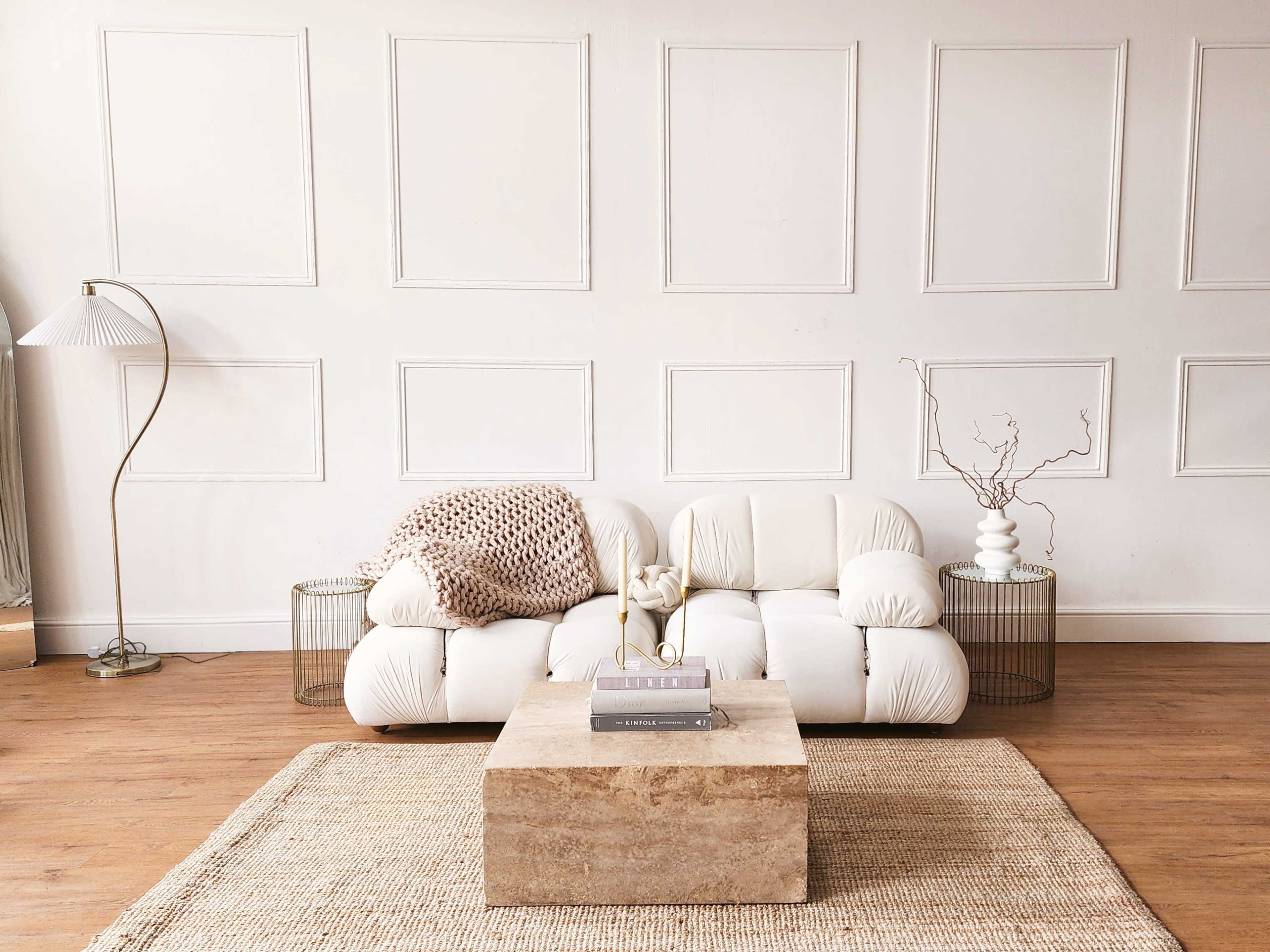 A modern living room featuring a light-colored sofa with a textured throw blanket, a wooden coffee table, and a floor lamp, against a wall with paneling.