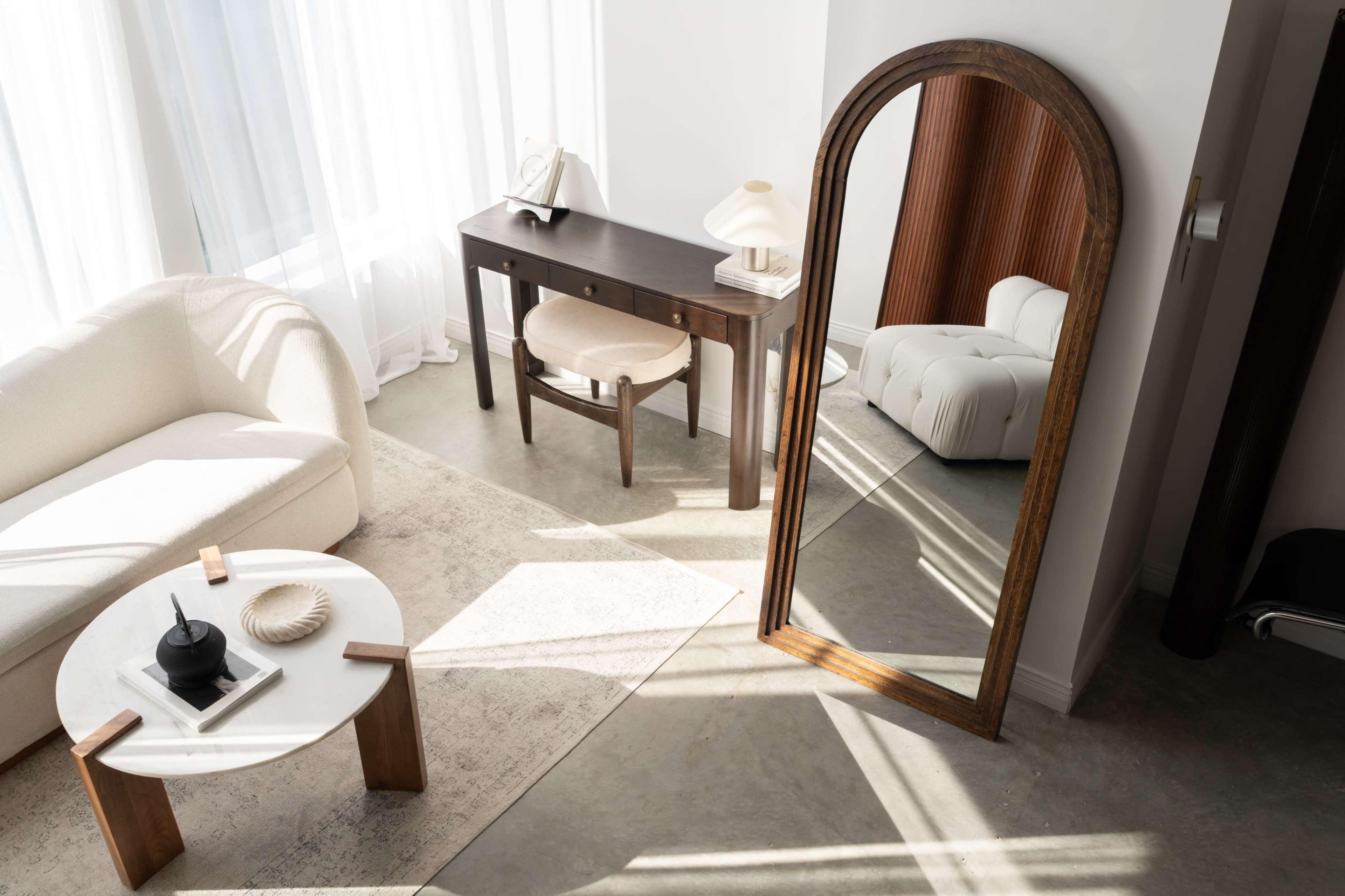 The image shows a bright, modern living area featuring a light-colored sofa, a round coffee table, and a large mirror leaning against the wall.