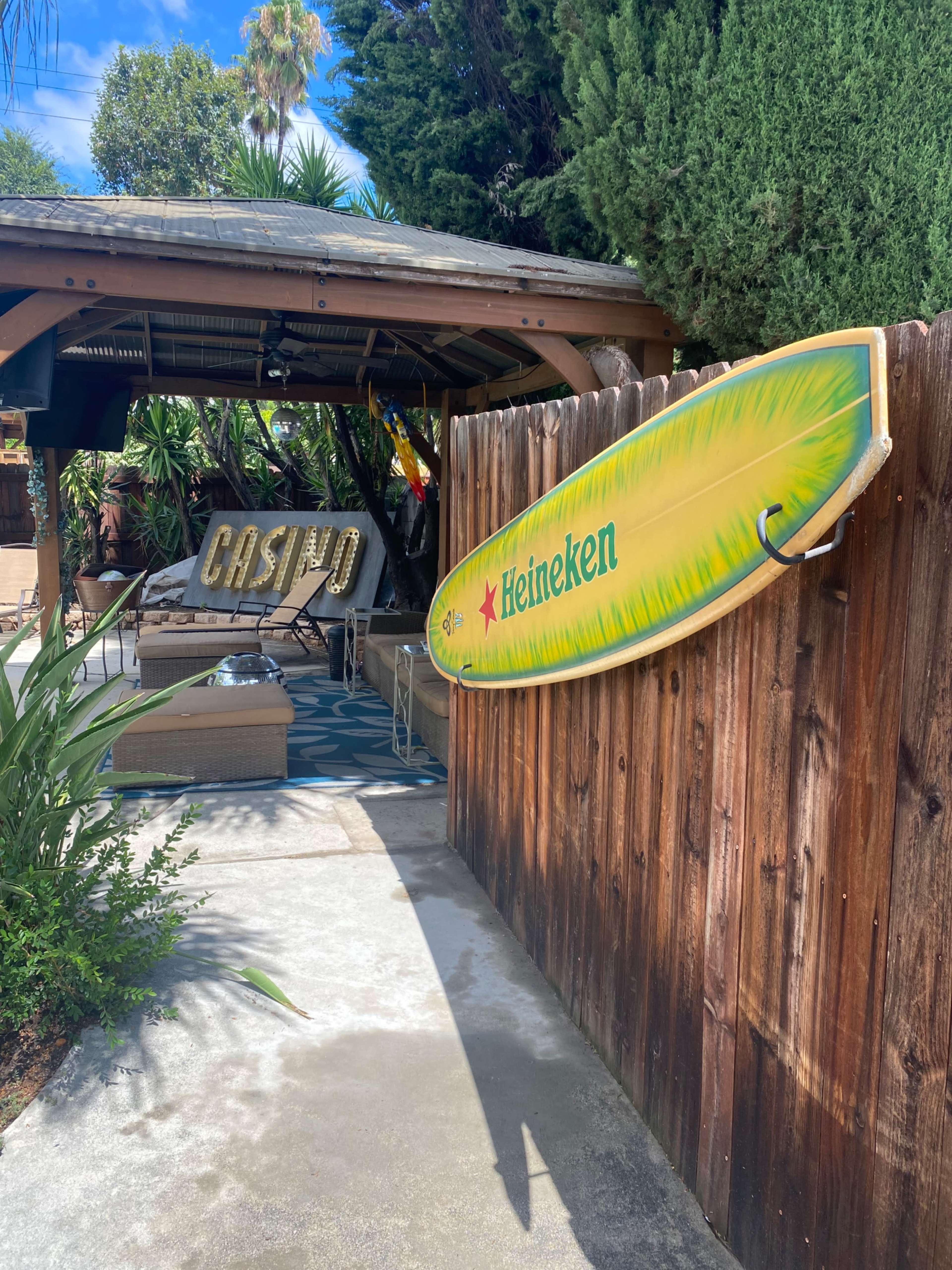 A wooden fence features a colorful surfboard sign with the Heineken logo near a shaded outdoor sitting area.