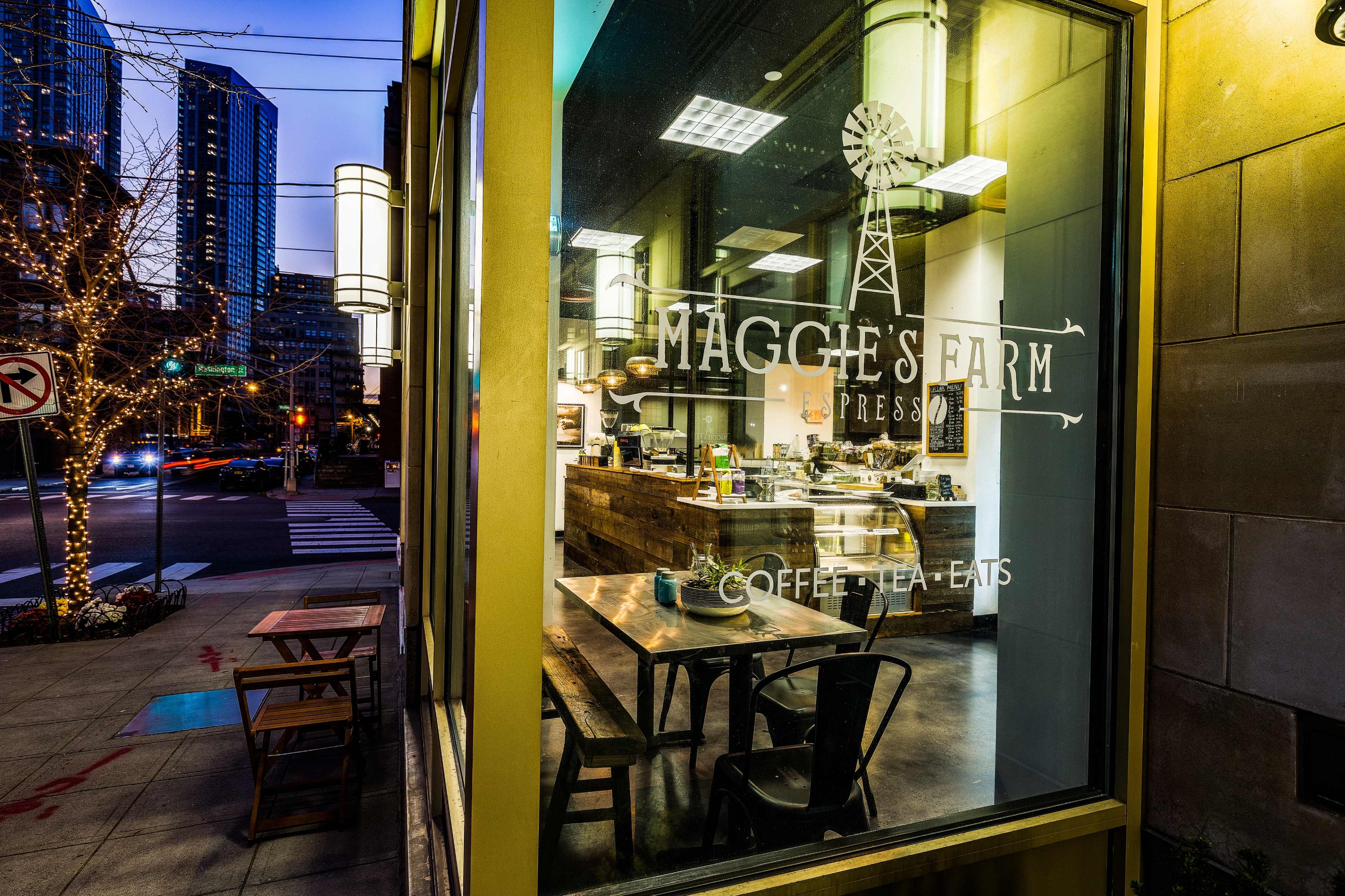 The exterior of Maggie's Farm coffee shop, featuring illuminated signage and a view of its wooden interior with tables and decor, set against a backdrop of city buildings at dusk.