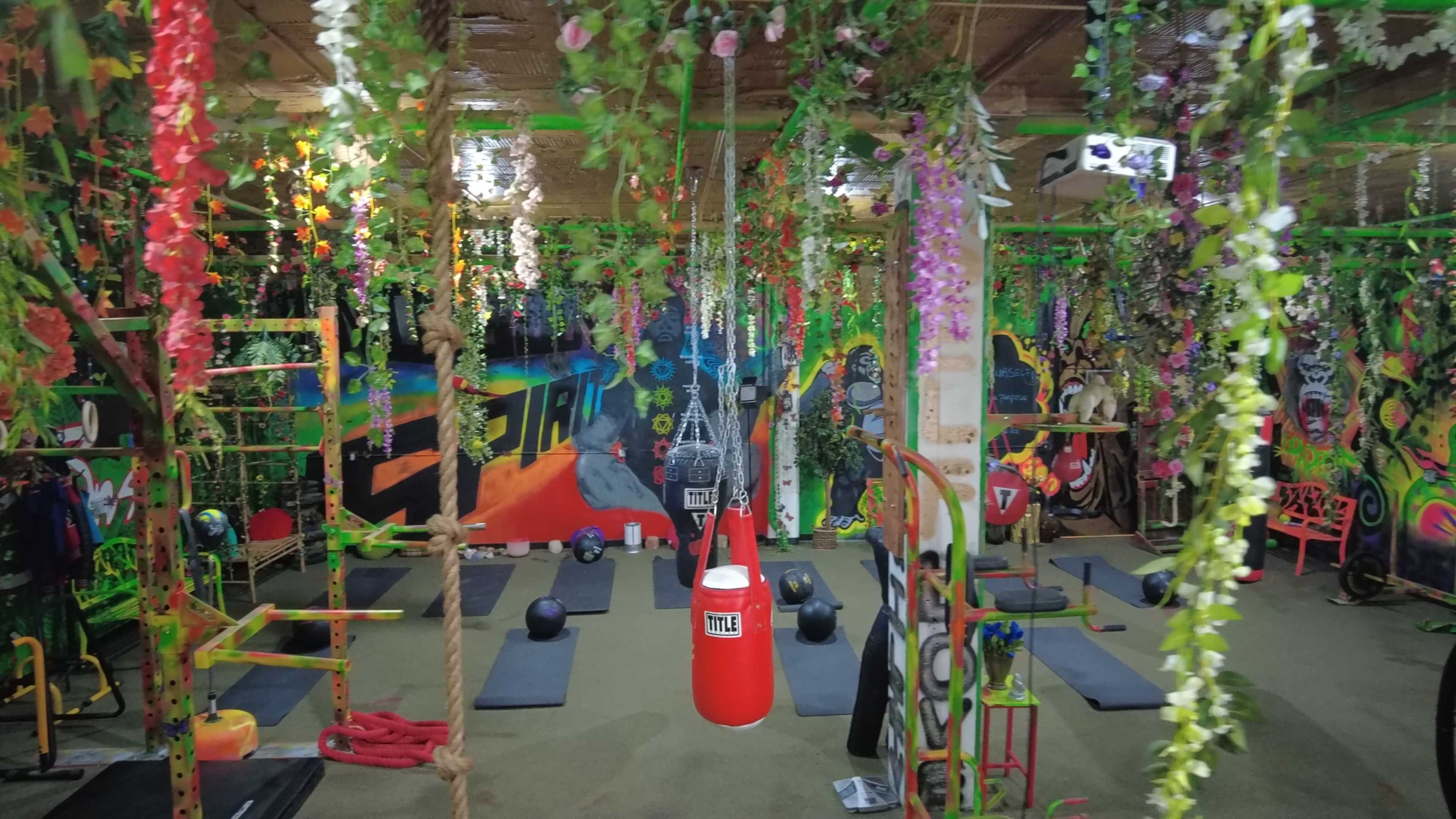 A colorful gym interior adorned with hanging plants and flowers, featuring exercise mats, a punching bag, and various workout equipment.