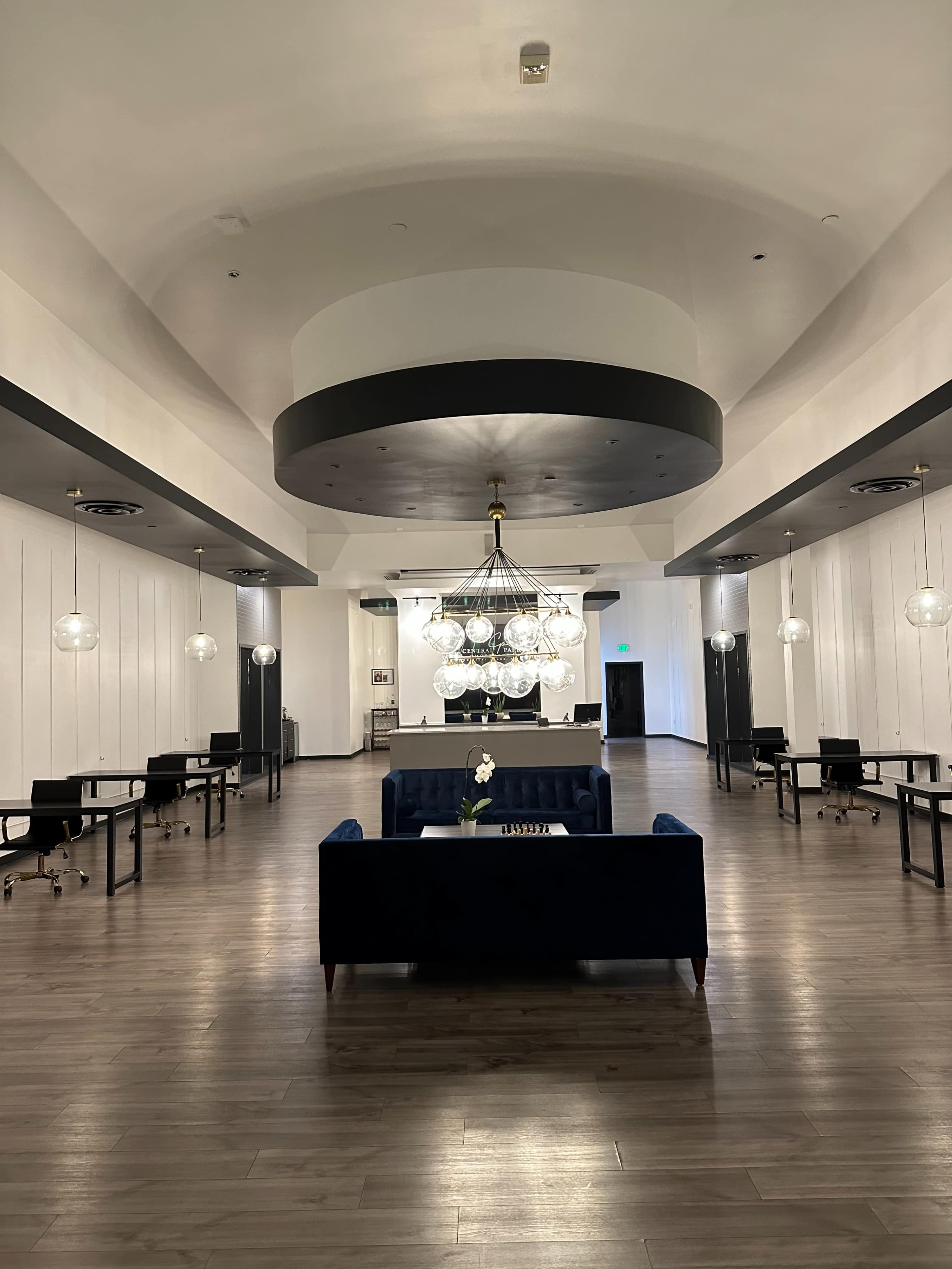 A spacious, modern interior with a central chandelier, seating area, and multiple workstations arranged symmetrically.