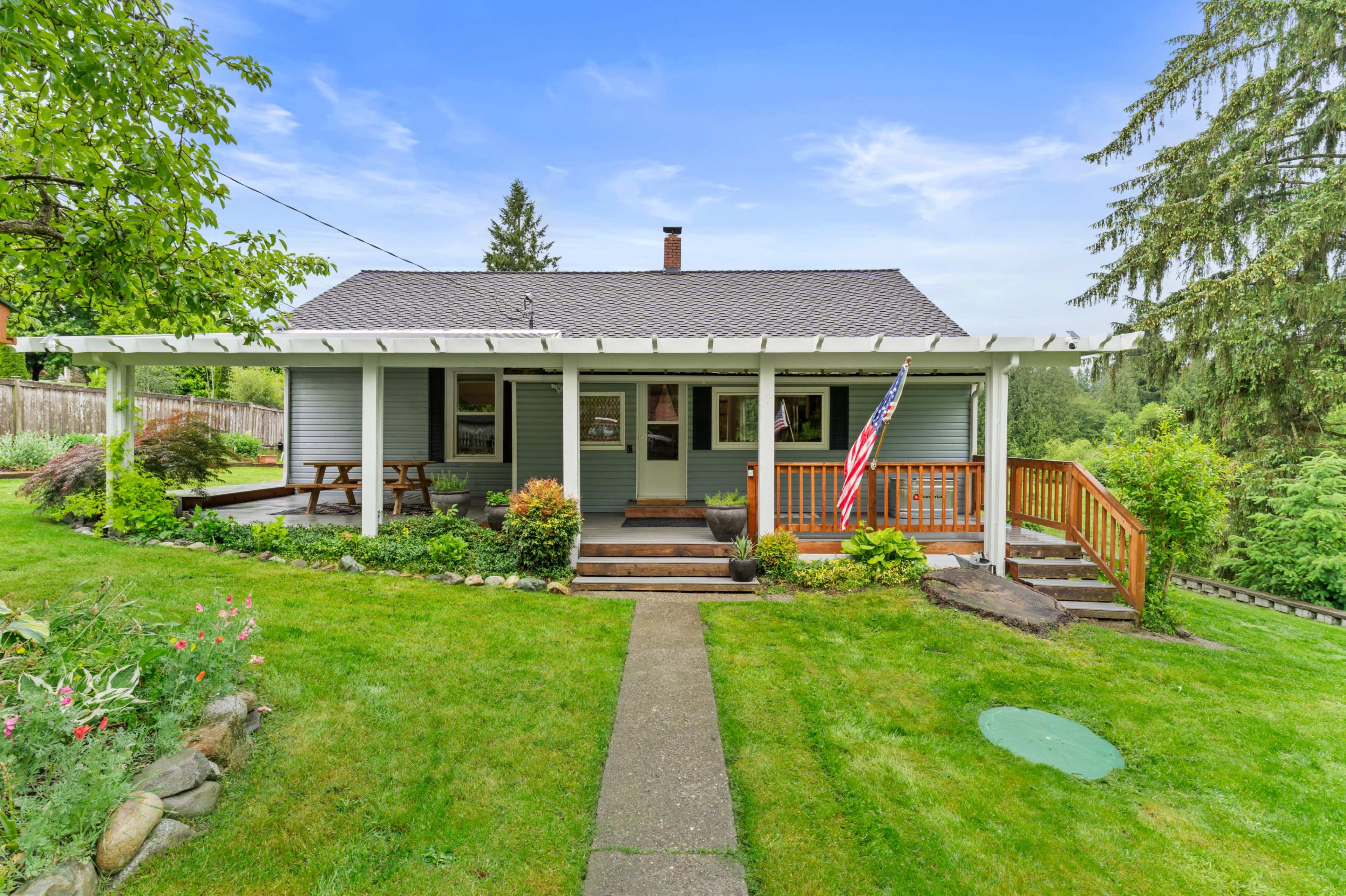 The image shows a charming single-story house with a front porch, surrounded by greenery and a well-maintained lawn.