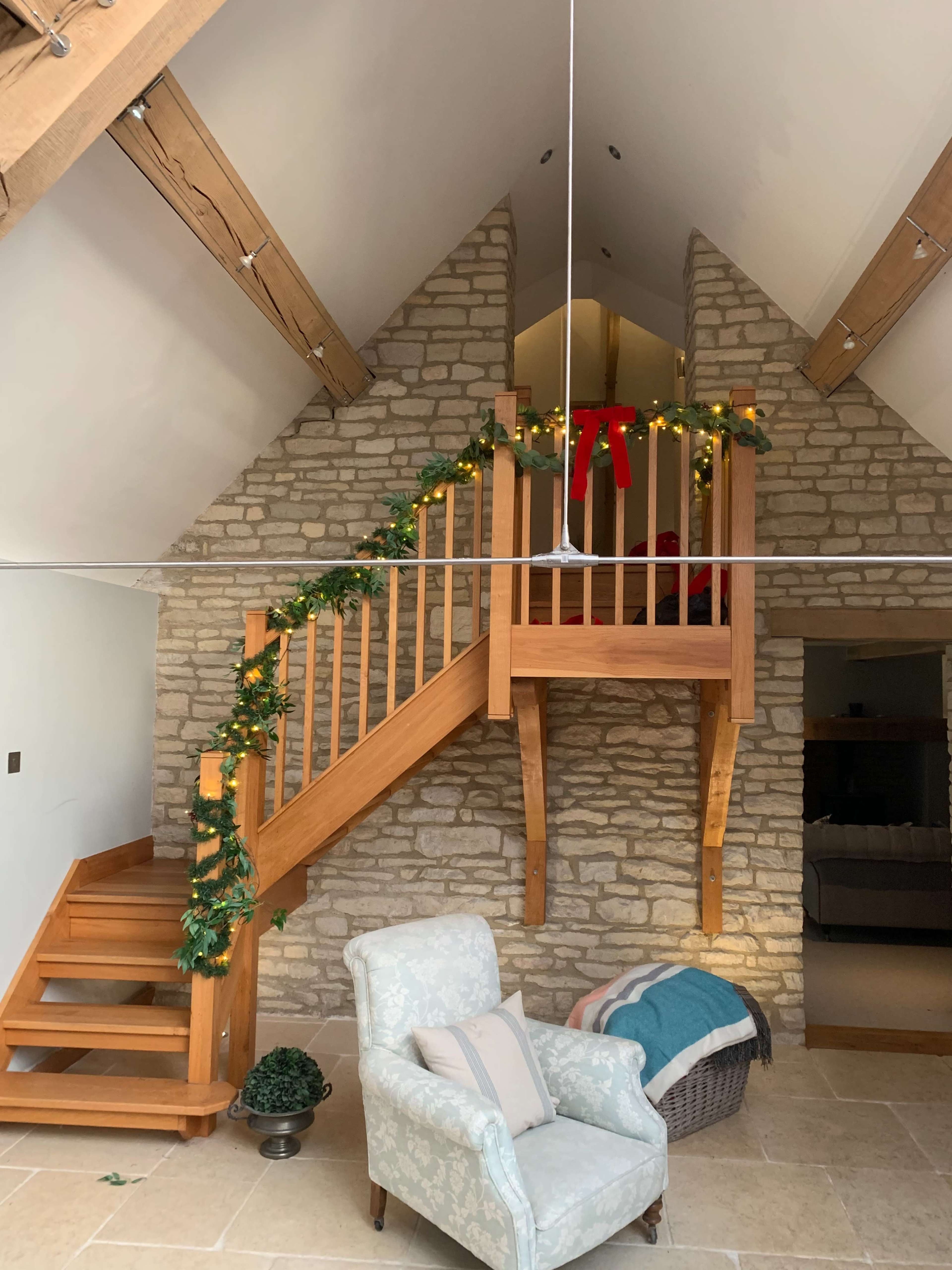A cozy interior space with a wooden staircase leading to a second level, adorned with festive greenery and a red bow, surrounded by stone walls and a light-colored armchair.