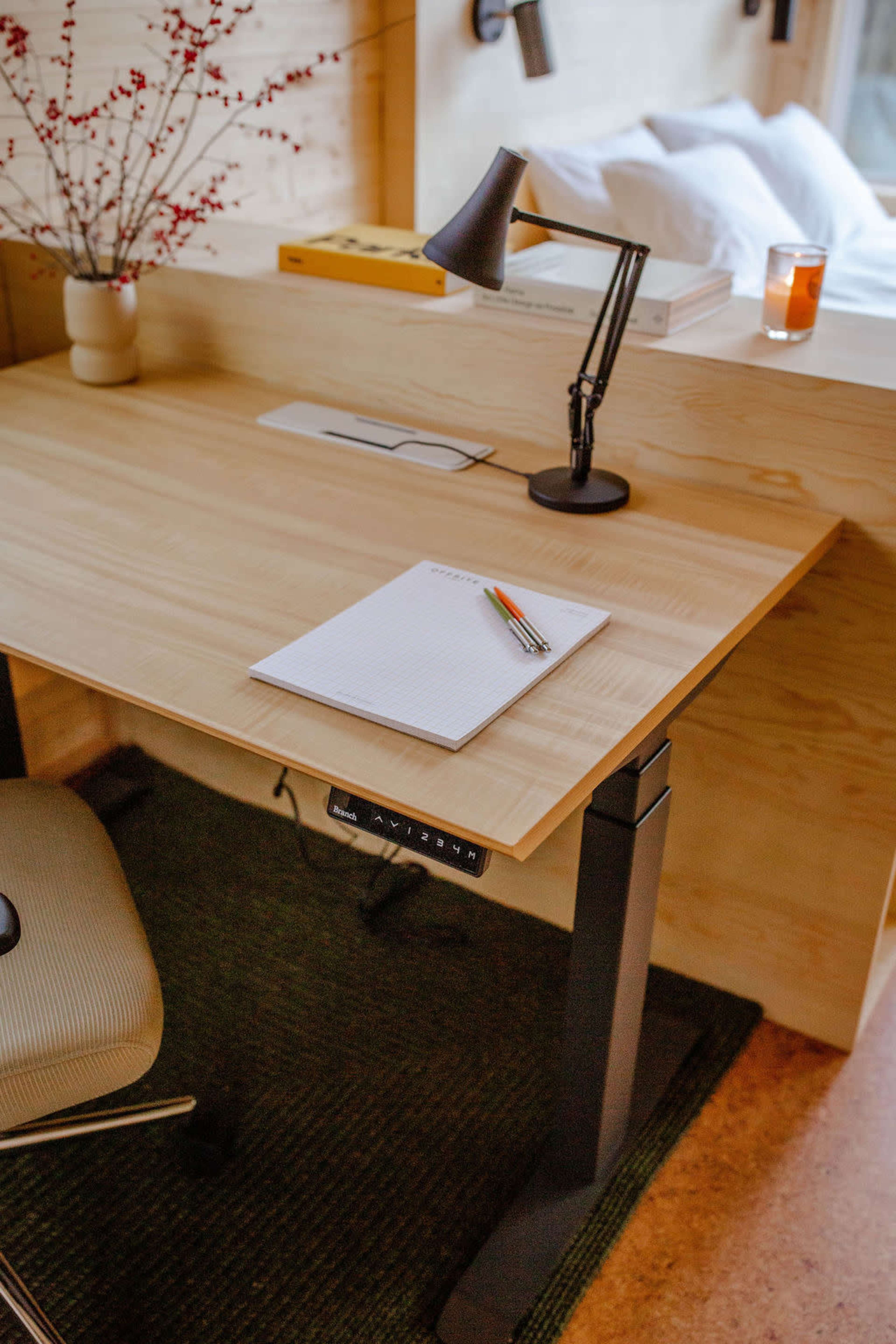 A minimalist wooden desk with a lamp, a notepad, colored pencils, and a small vase of branches, positioned near a window.