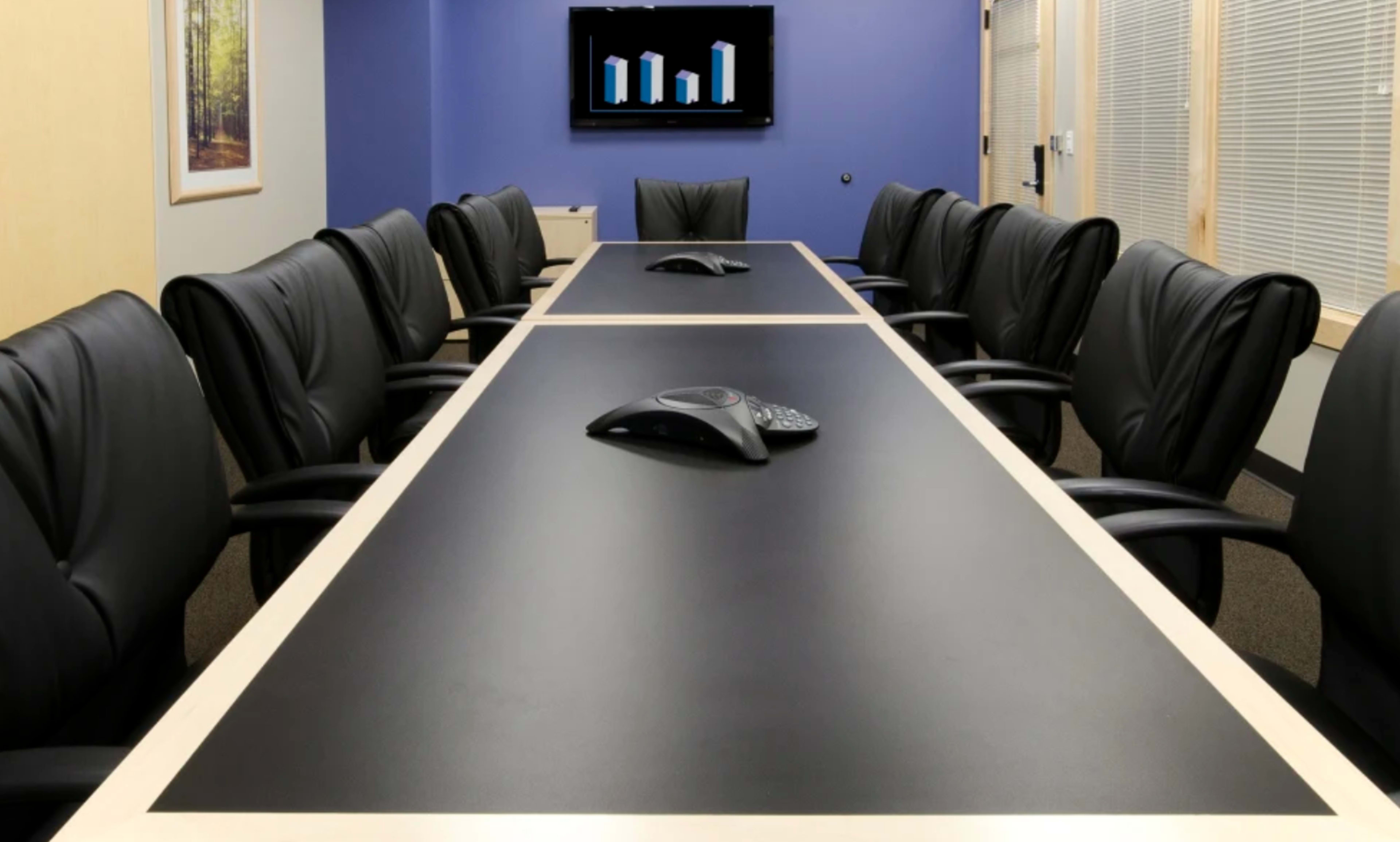 A conference room features a long table with black leather chairs and a screen displaying bar graphs on a blue wall.