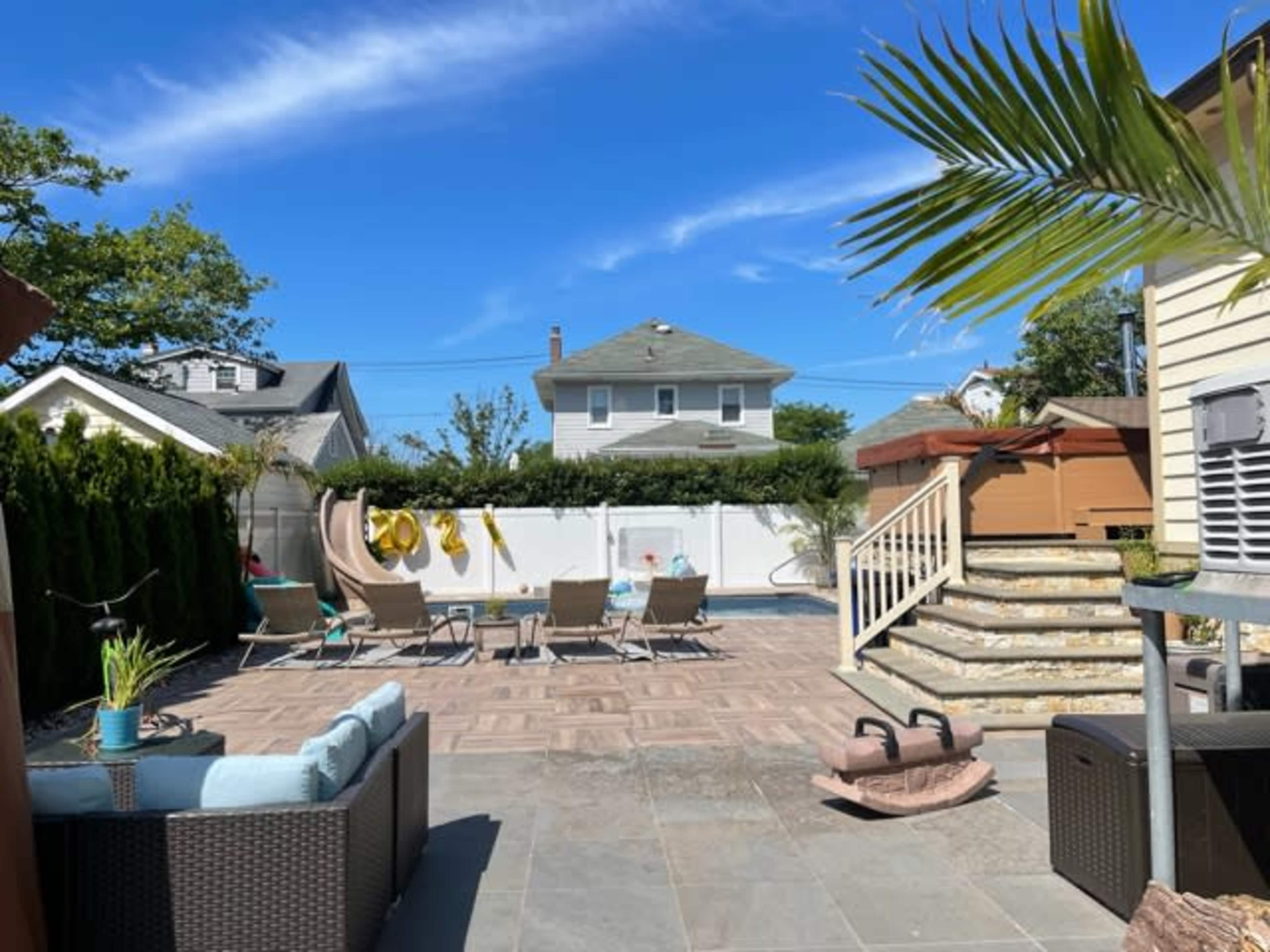 The image shows a backyard with a swimming pool, lounging chairs, a slide, and a deck area under a clear blue sky.