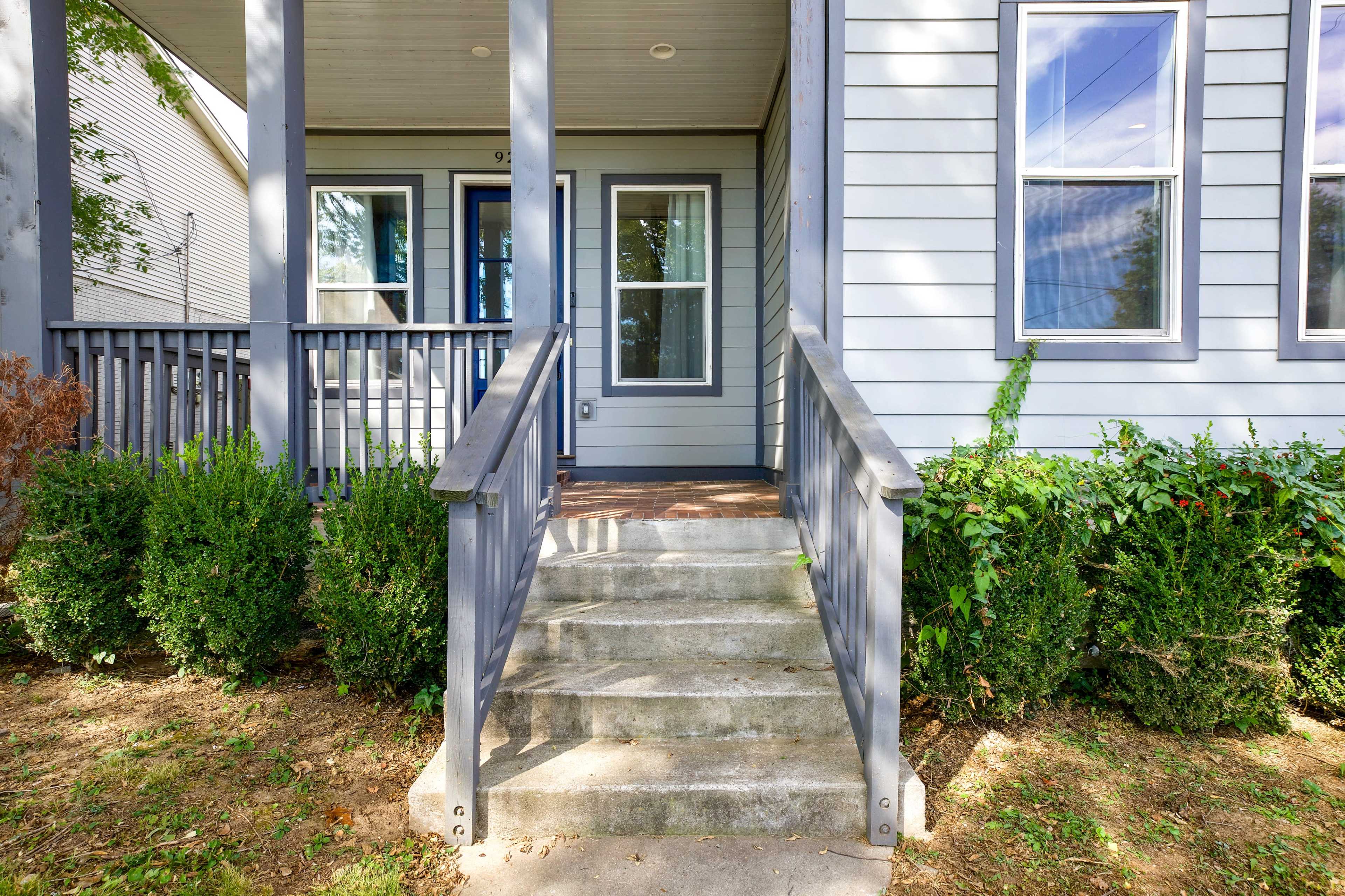 A set of concrete steps leading to a front porch with a blue railing and two doors on a gray house.