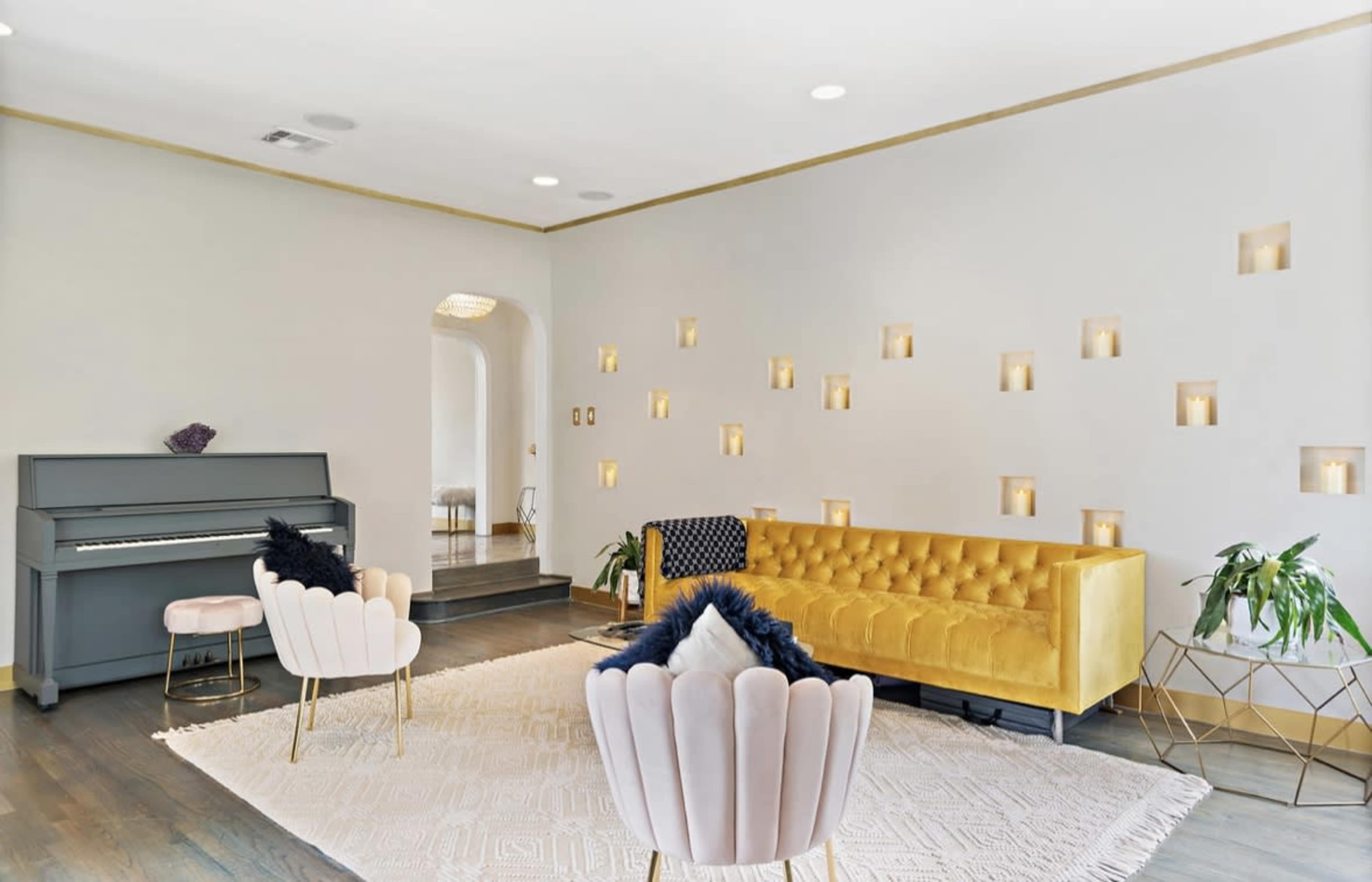 A modern living room with a yellow tufted sofa, a gray piano, two pink chairs, and wall niches illuminated with soft lighting.