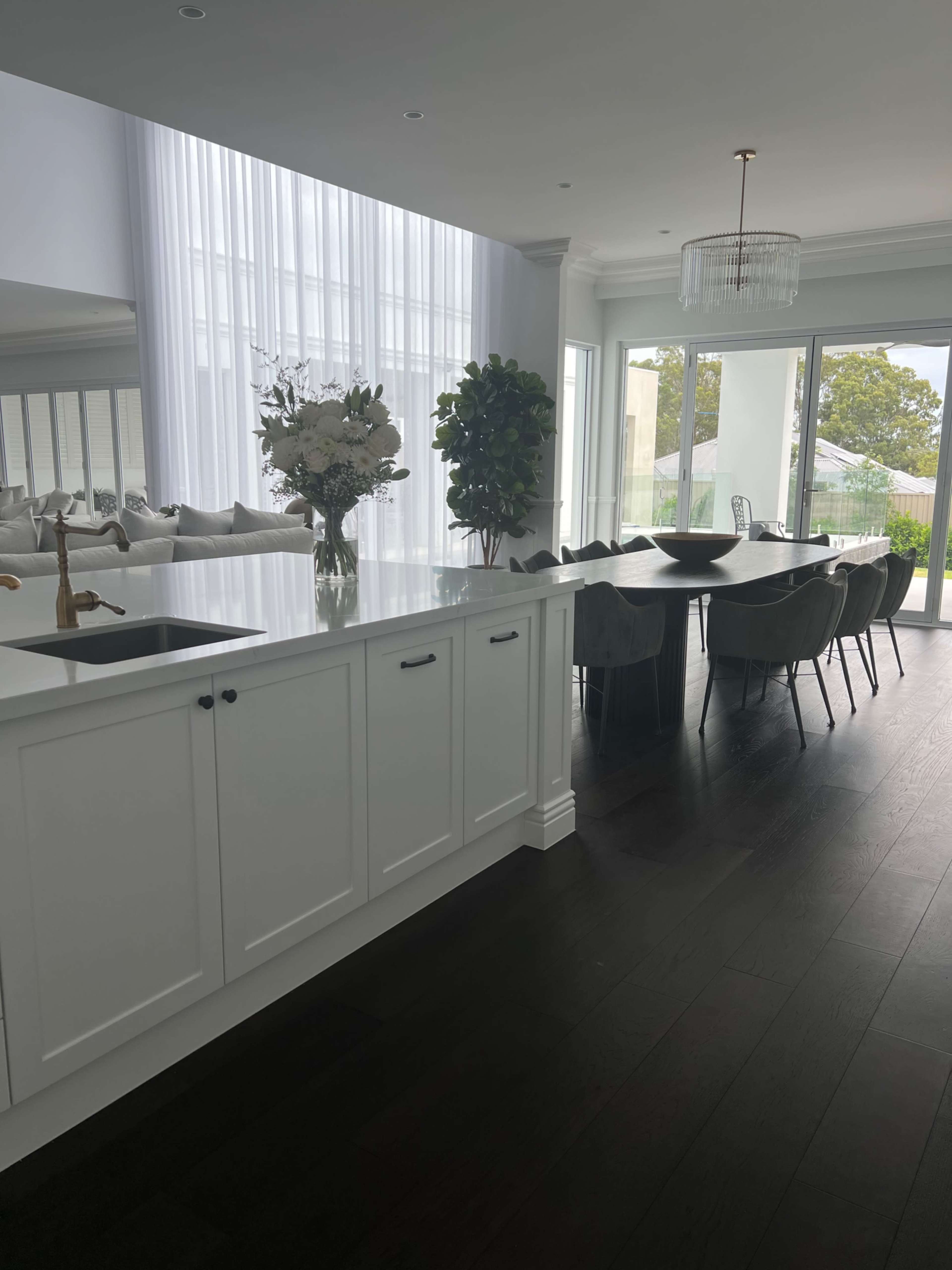 The image shows a modern kitchen featuring a white island with a sink, a dining area with a long table and upholstered chairs, and large windows allowing natural light to enter.