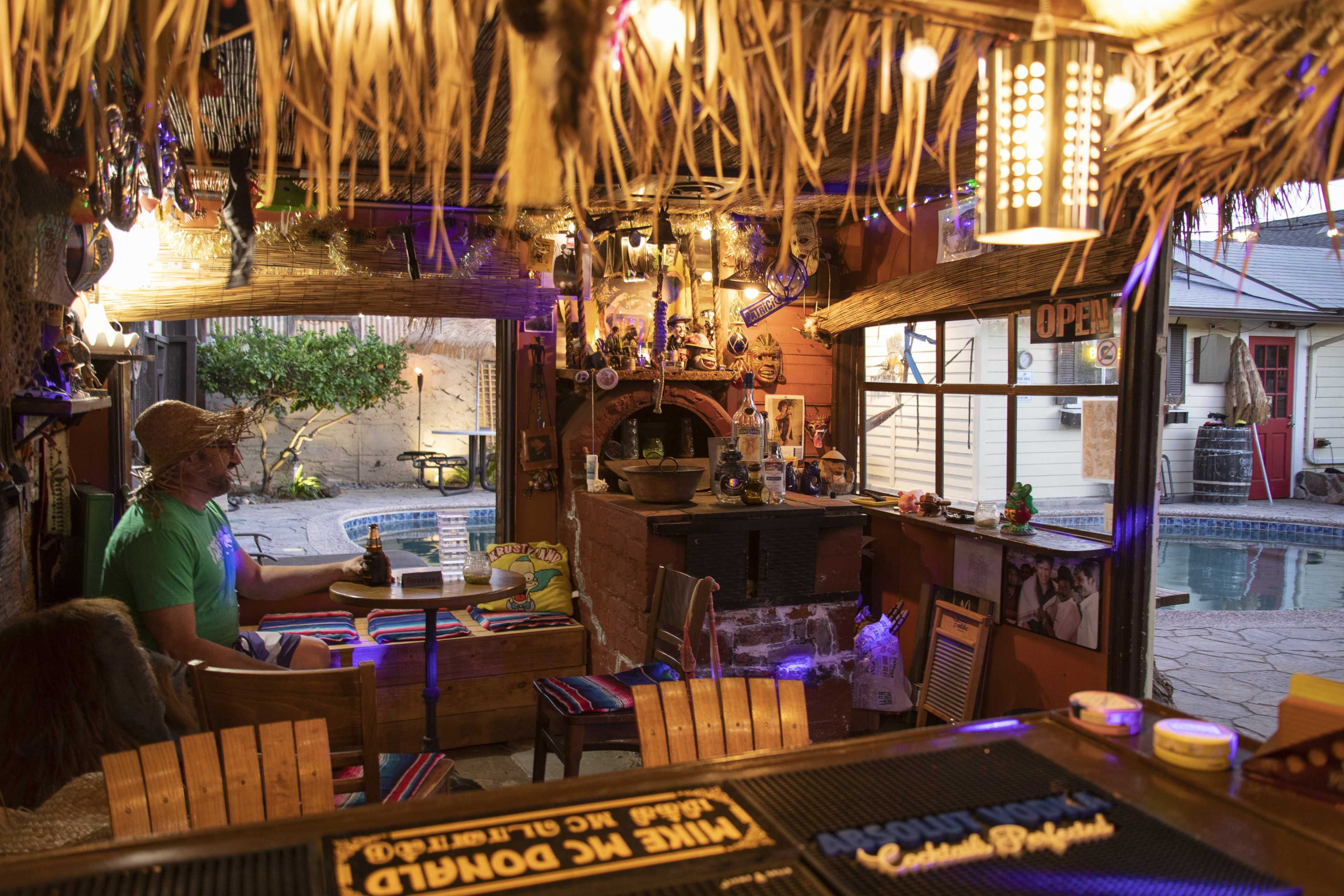A person sits at a rustic bar inside a thatched-roof tiki lounge, with a pool and courtyard visible through large windows.