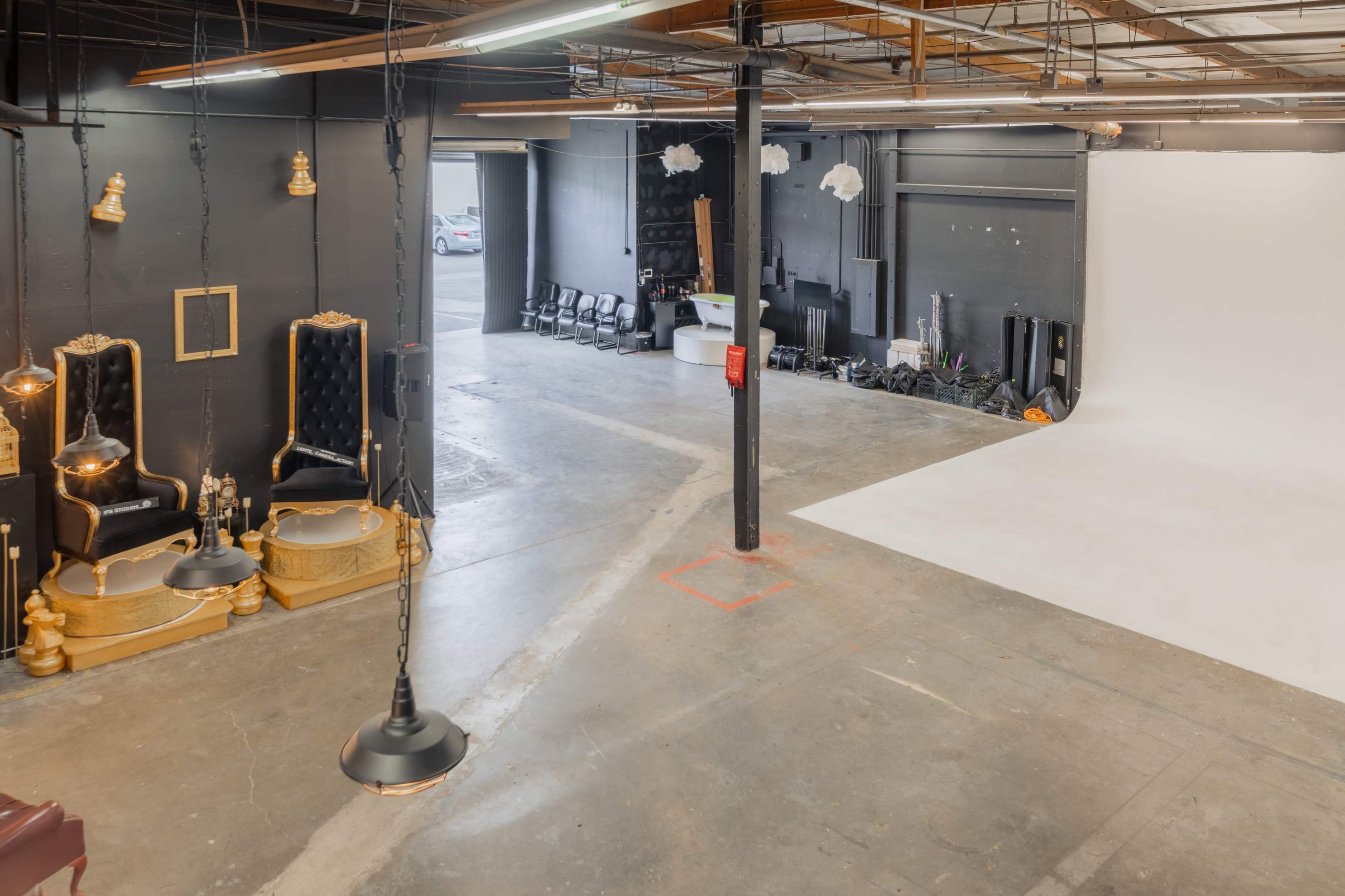 The image shows a spacious studio with a white backdrop, black walls, and decorative chairs on one side, while equipment and chairs are arranged in the background.