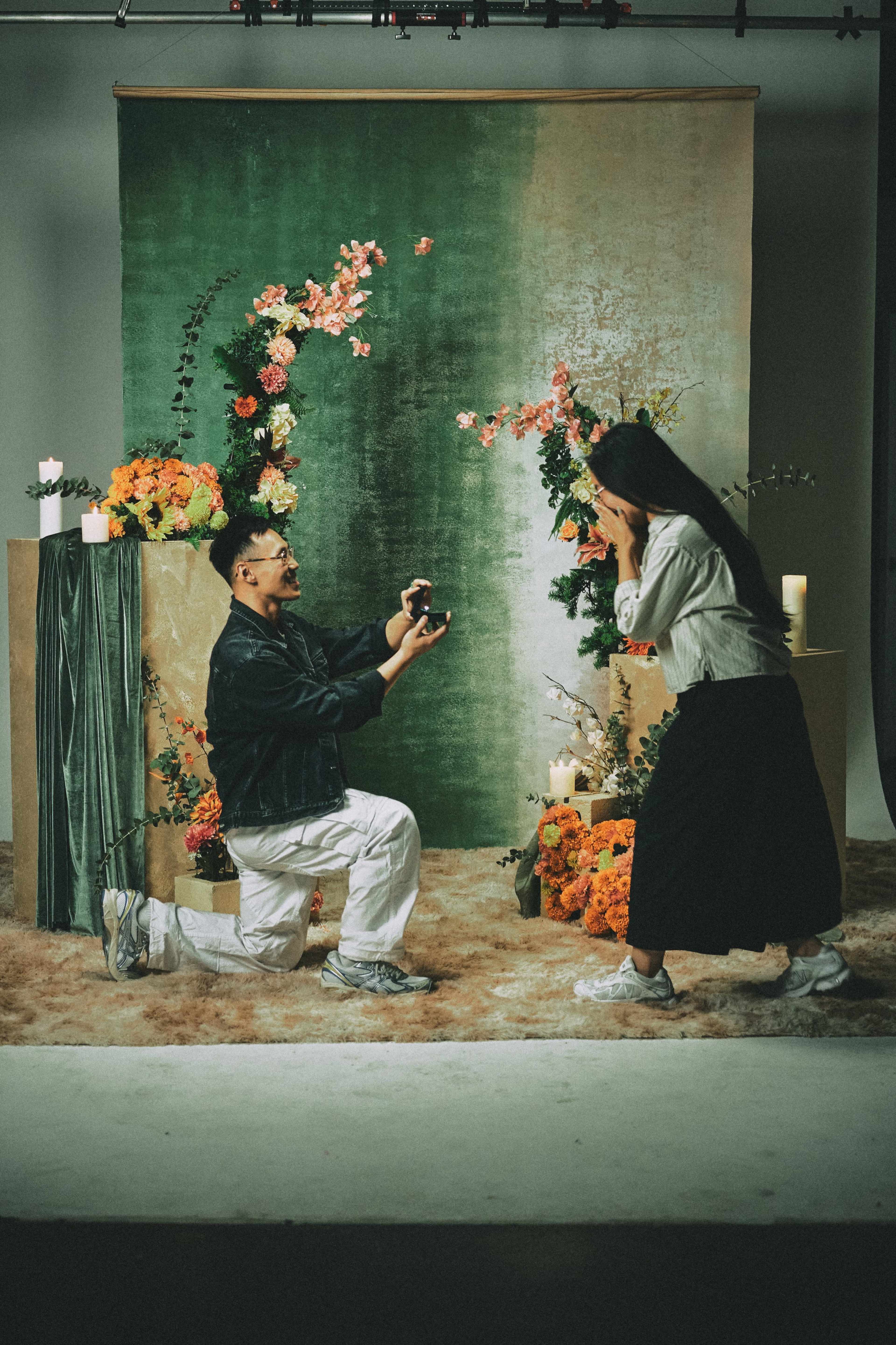 A man kneels with a ring in a proposal, while a woman stands nearby with her hands covering her face, surrounded by floral decorations.