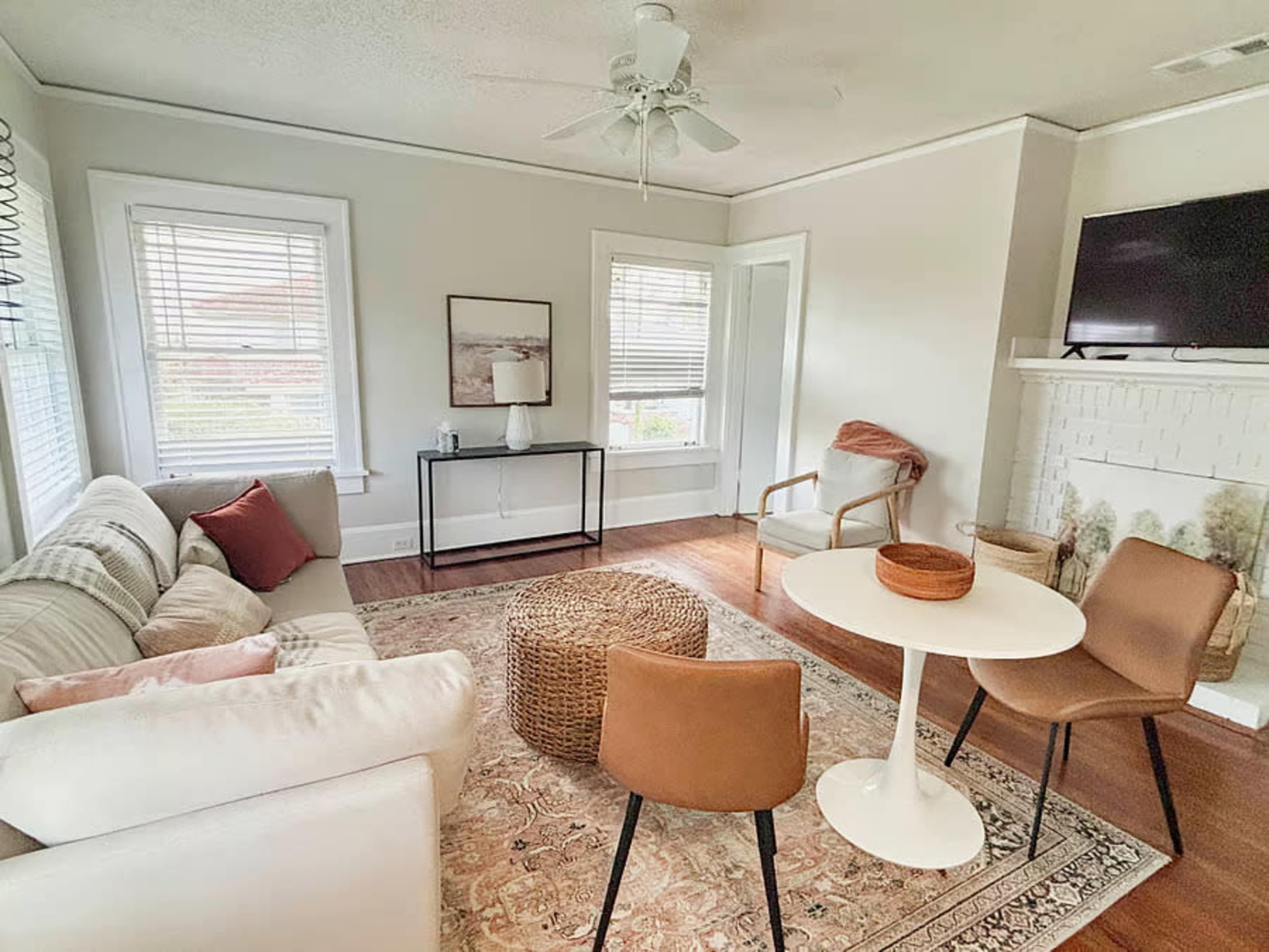 The image shows a cozy living room with a white sofa, a round table, two brown chairs, and a woven ottoman on a patterned rug.
