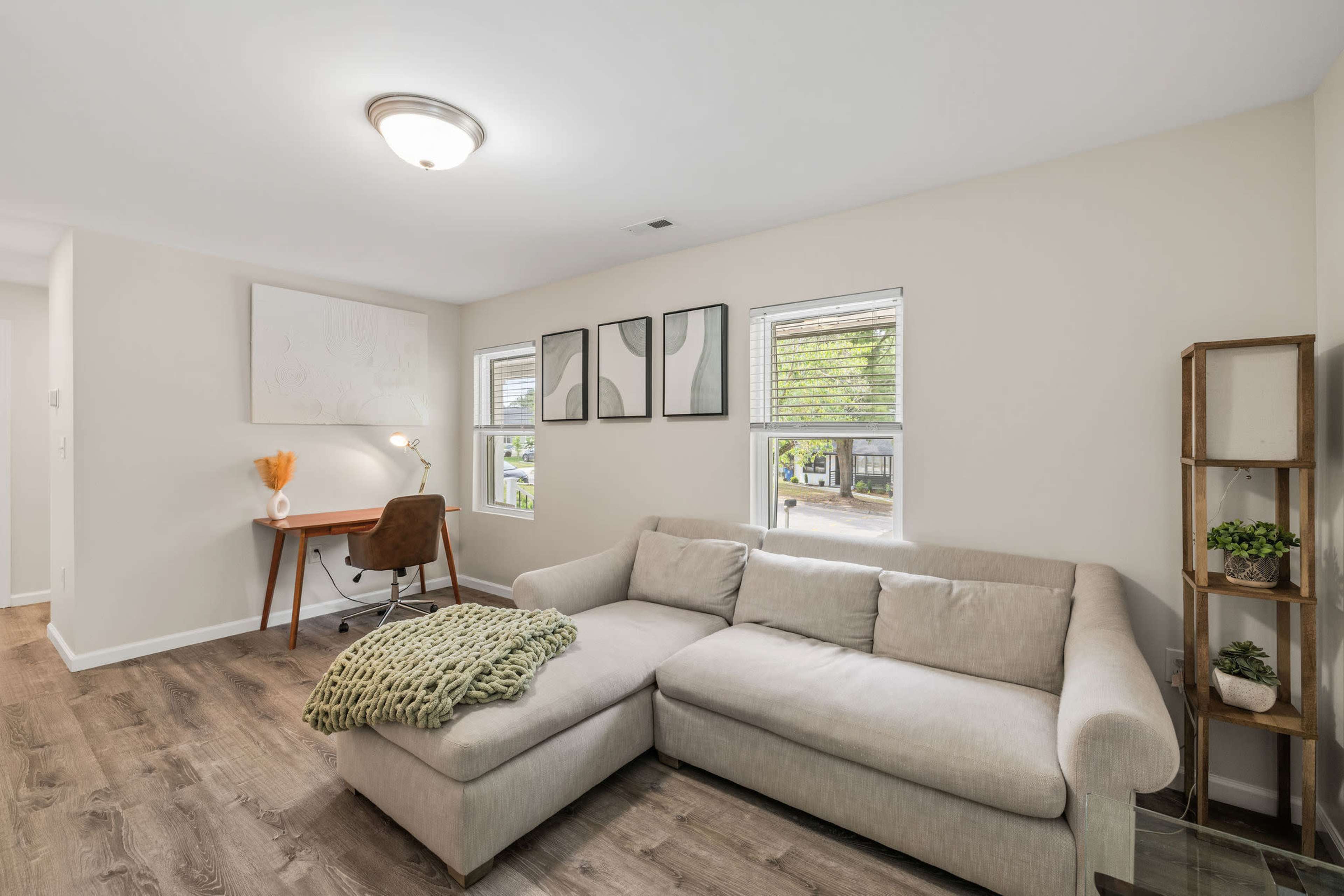 A modern living room features a light-colored sectional sofa, a wooden desk with a chair, and framed artwork on the wall, complemented by large windows that allow natural light.