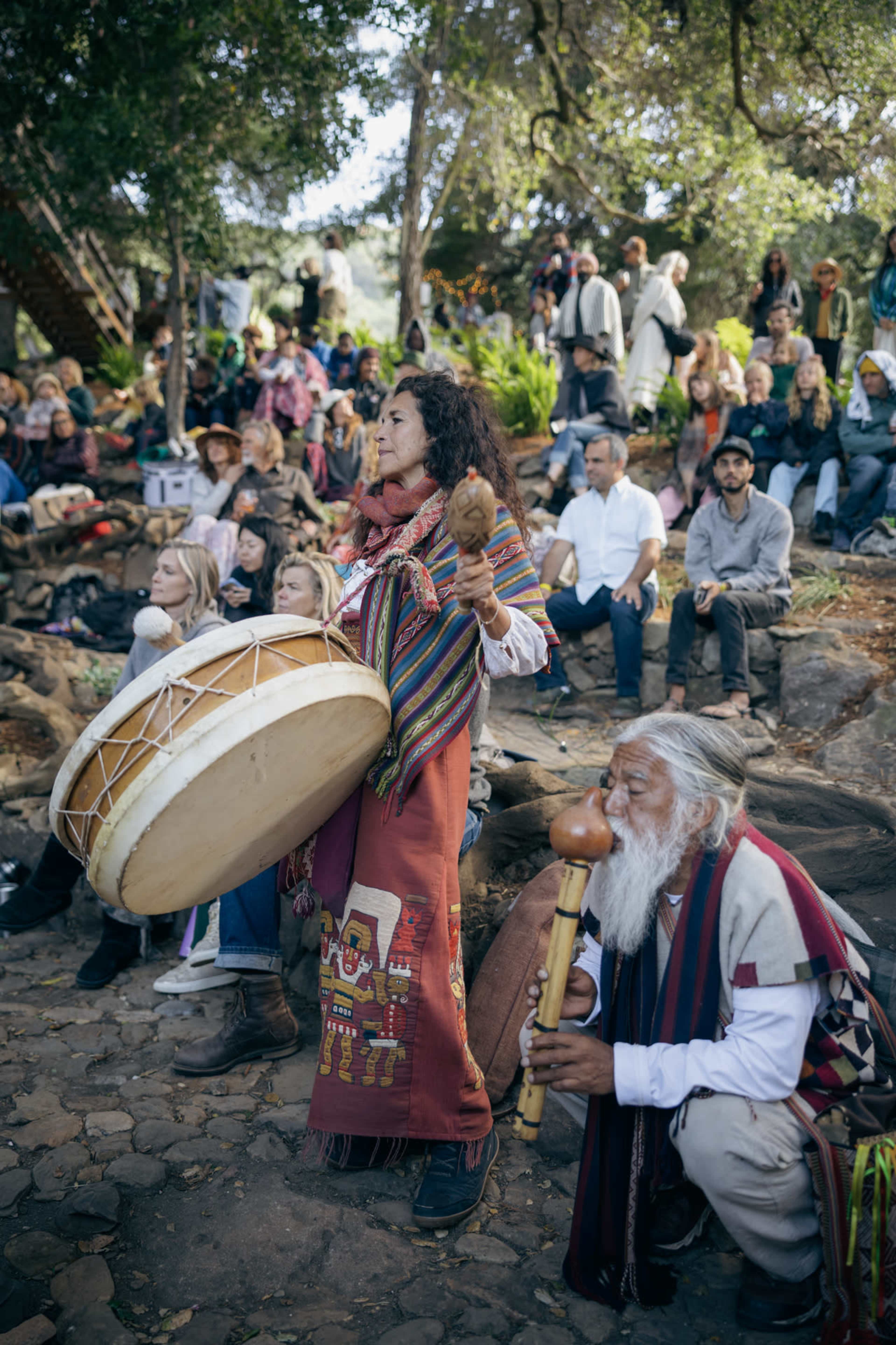 A woman in traditional attire plays a large drum while an elderly man with a gourd instrument sits nearby, surrounded by a diverse audience in an outdoor gathering.