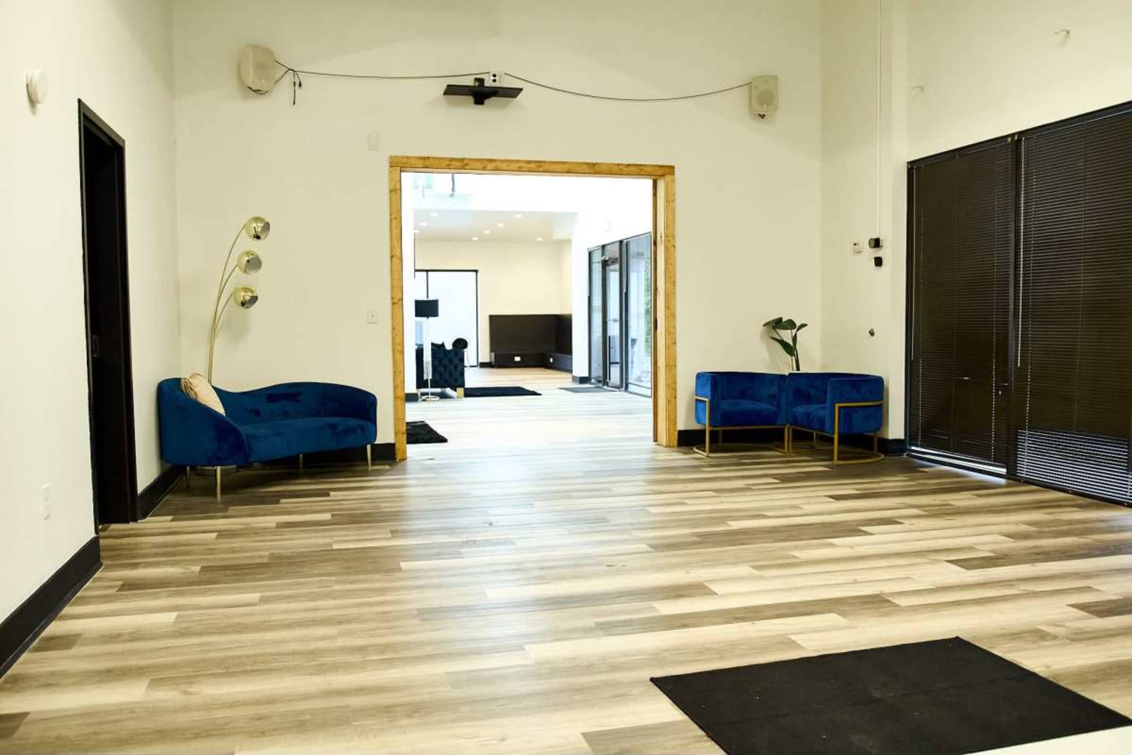 The image shows a spacious interior with wooden flooring, two blue velvet chairs on the left, and an open doorway leading to another area with a TV in the background.