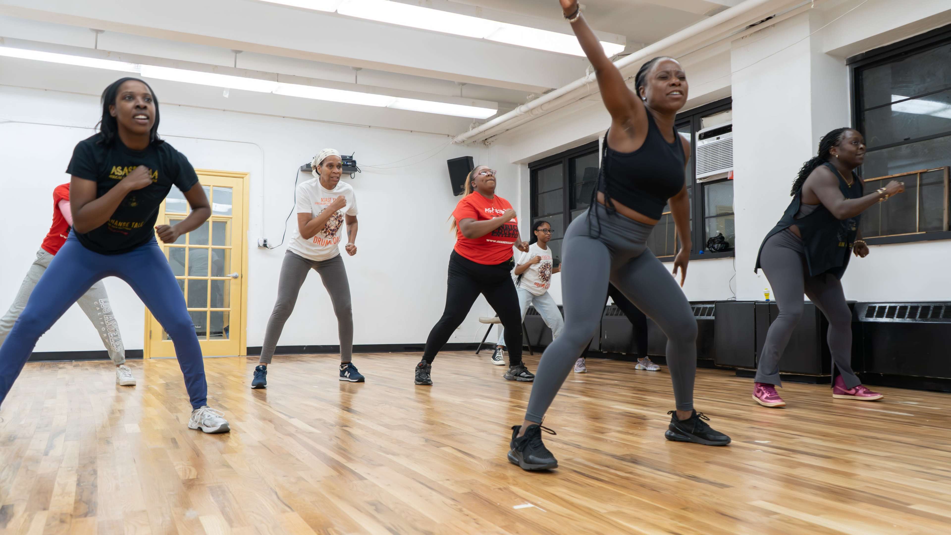 A group of seven women are engaged in a fitness class, demonstrating various movements in a bright, spacious studio with wooden floors.