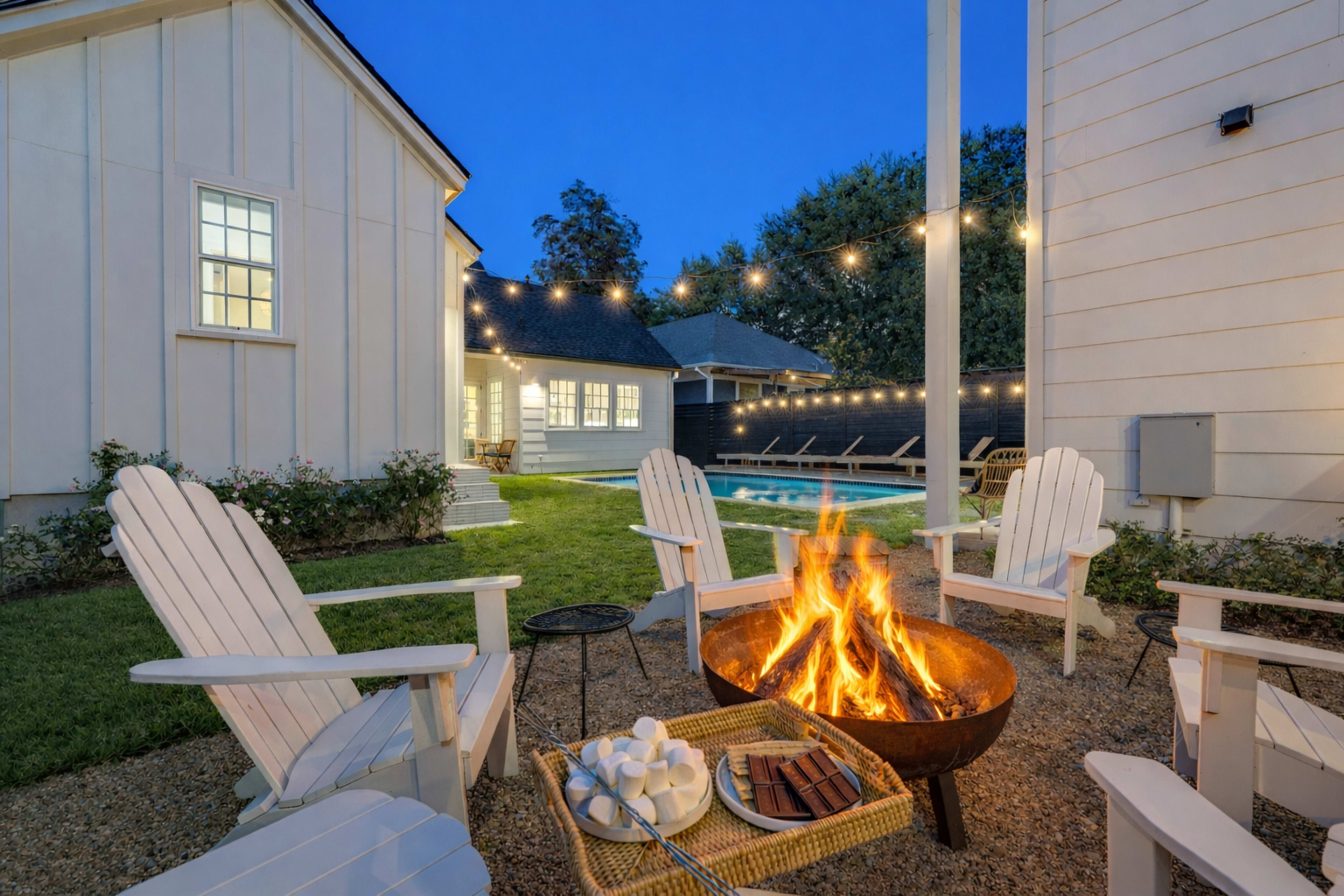 A cozy outdoor seating area features four white Adirondack chairs arranged around a fire pit, with a backyard pool and string lights in the background.