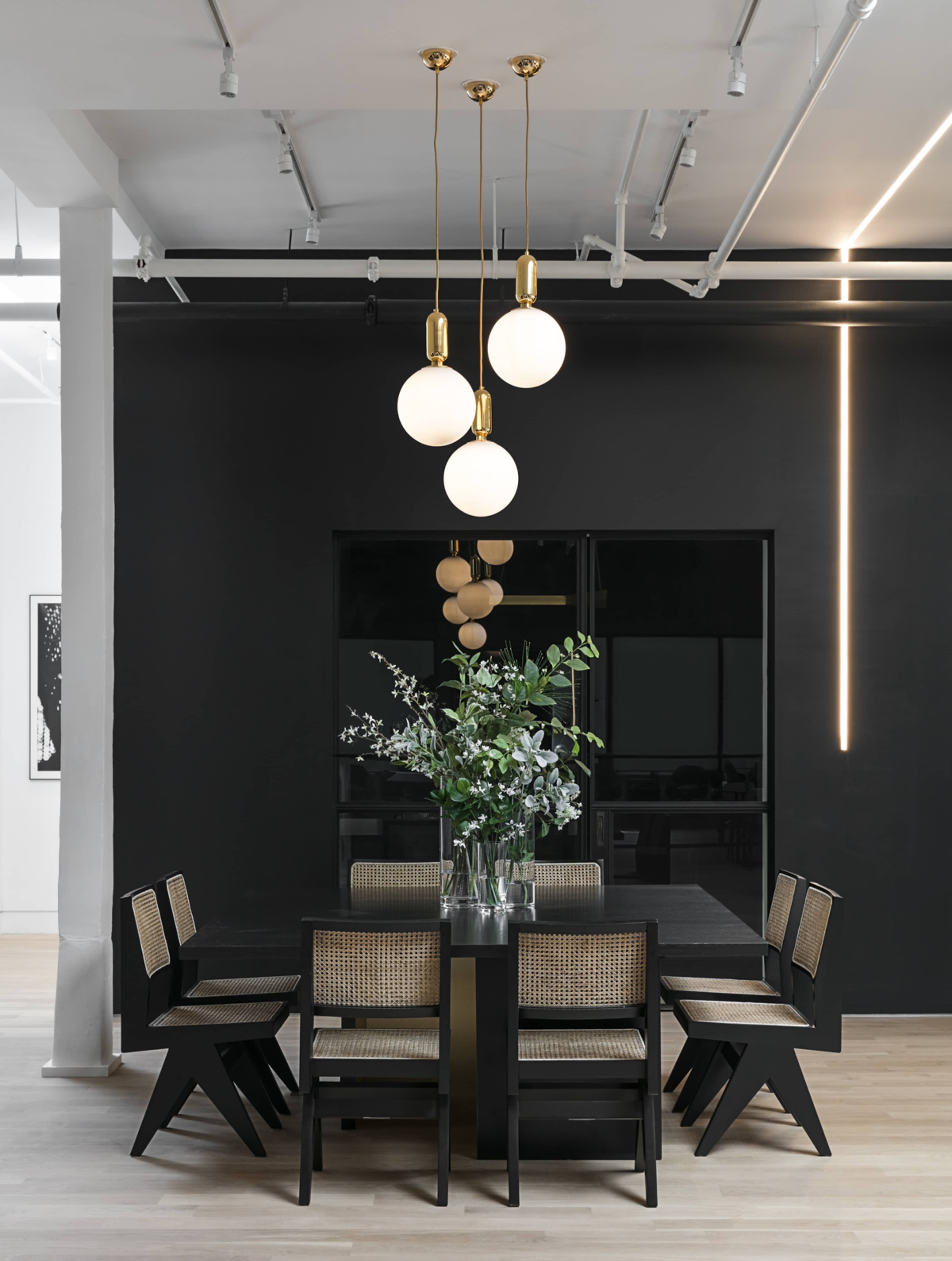 A minimalist dining area features a large black table surrounded by six chairs, with pendant lights hanging above and a vase of greenery as the centerpiece.