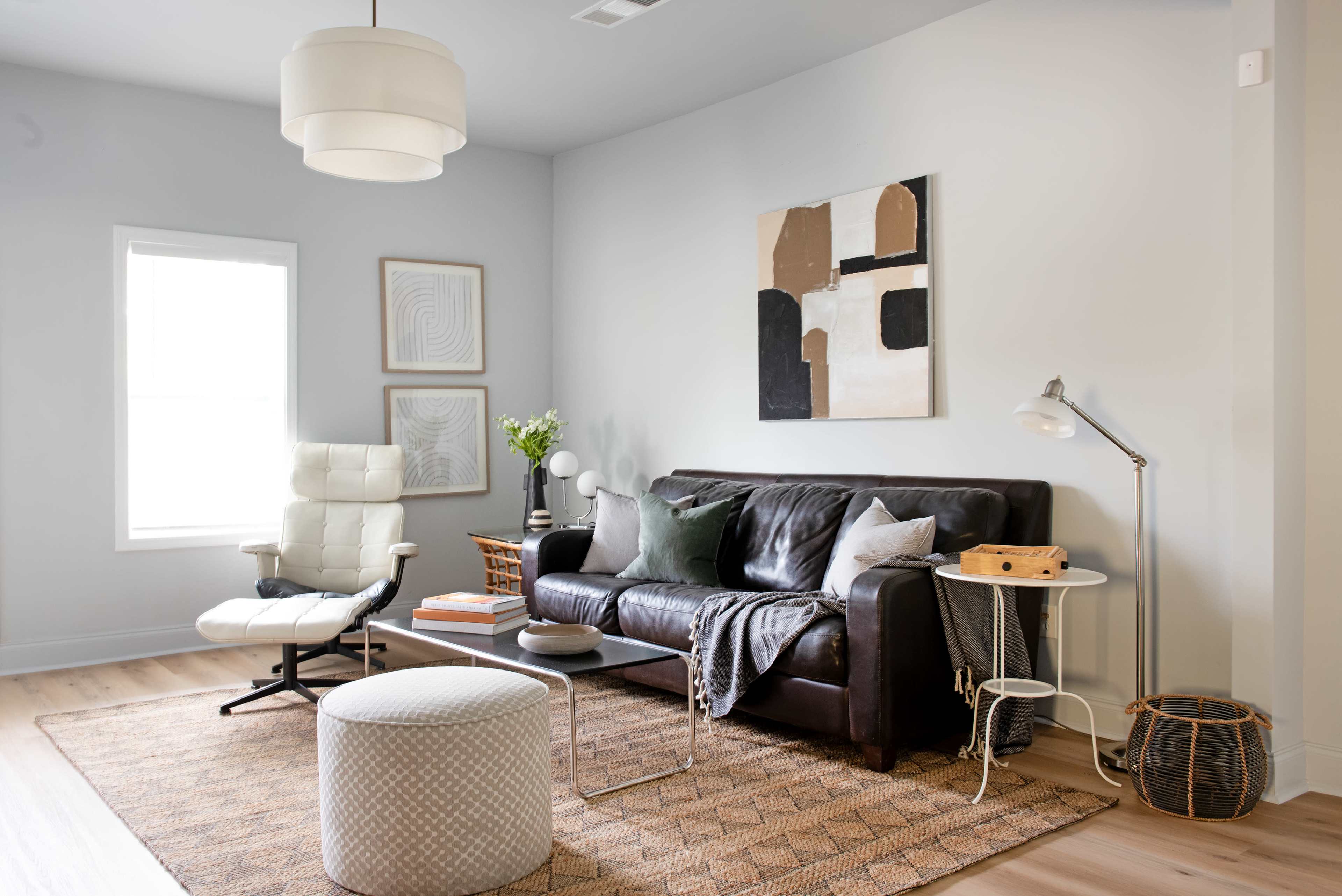 A modern living room features a dark leather couch, an armchair, a round ottoman, a patterned area rug, and a wall art piece, all complemented by natural light from a window.