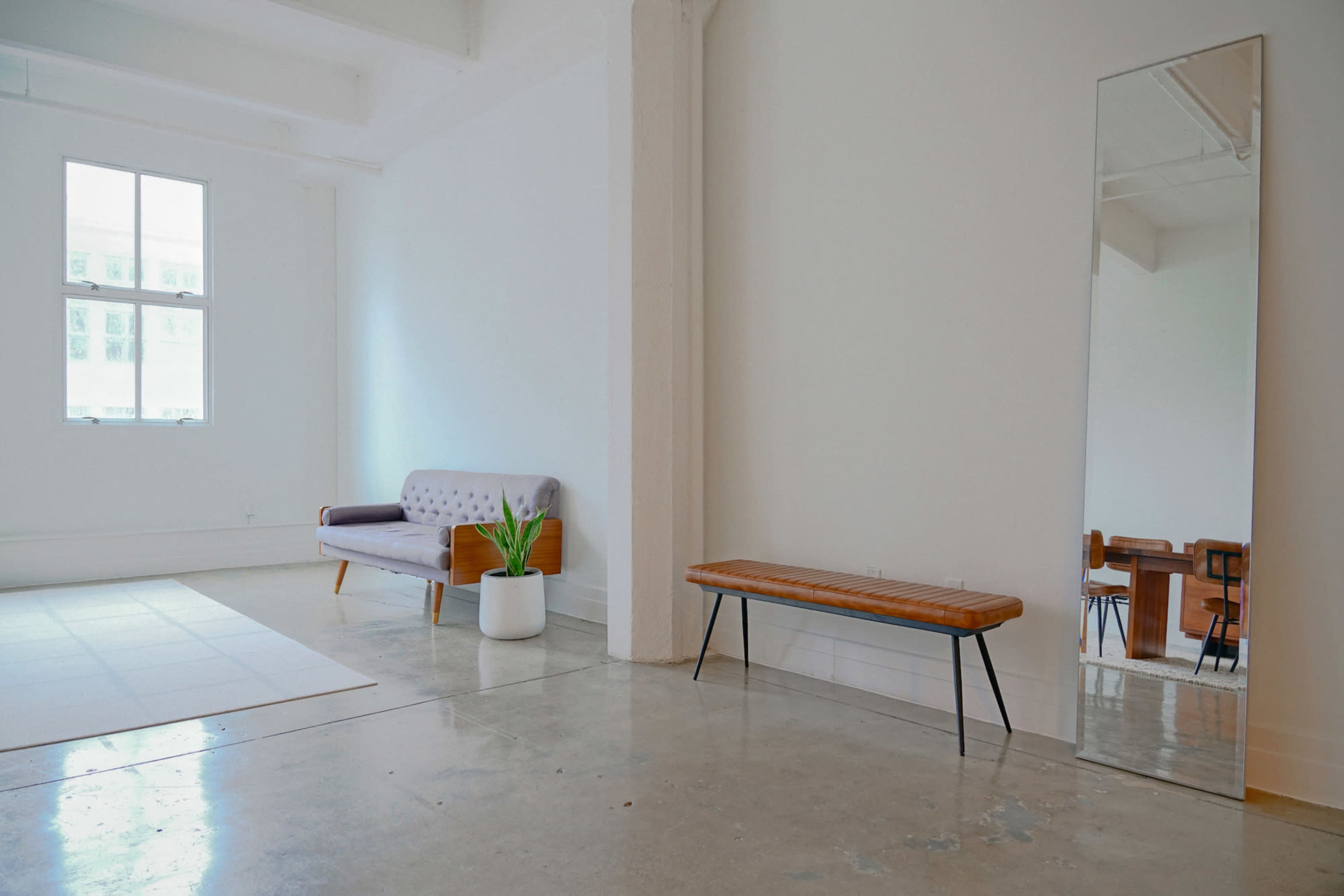 A minimalist room with a sofa, wooden bench, potted plant, and a large mirror against white walls and a polished concrete floor.