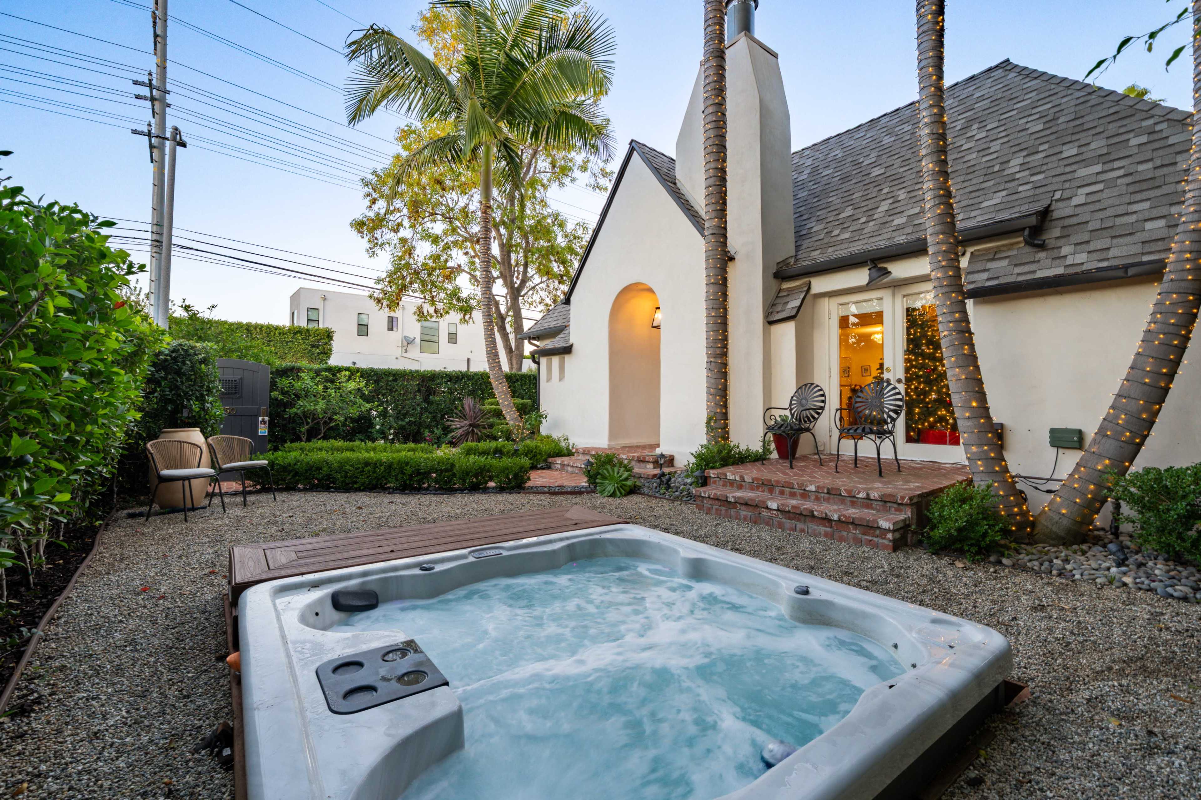 A hot tub is located on a gravel patio in a landscaped backyard with palm trees and a house in the background.