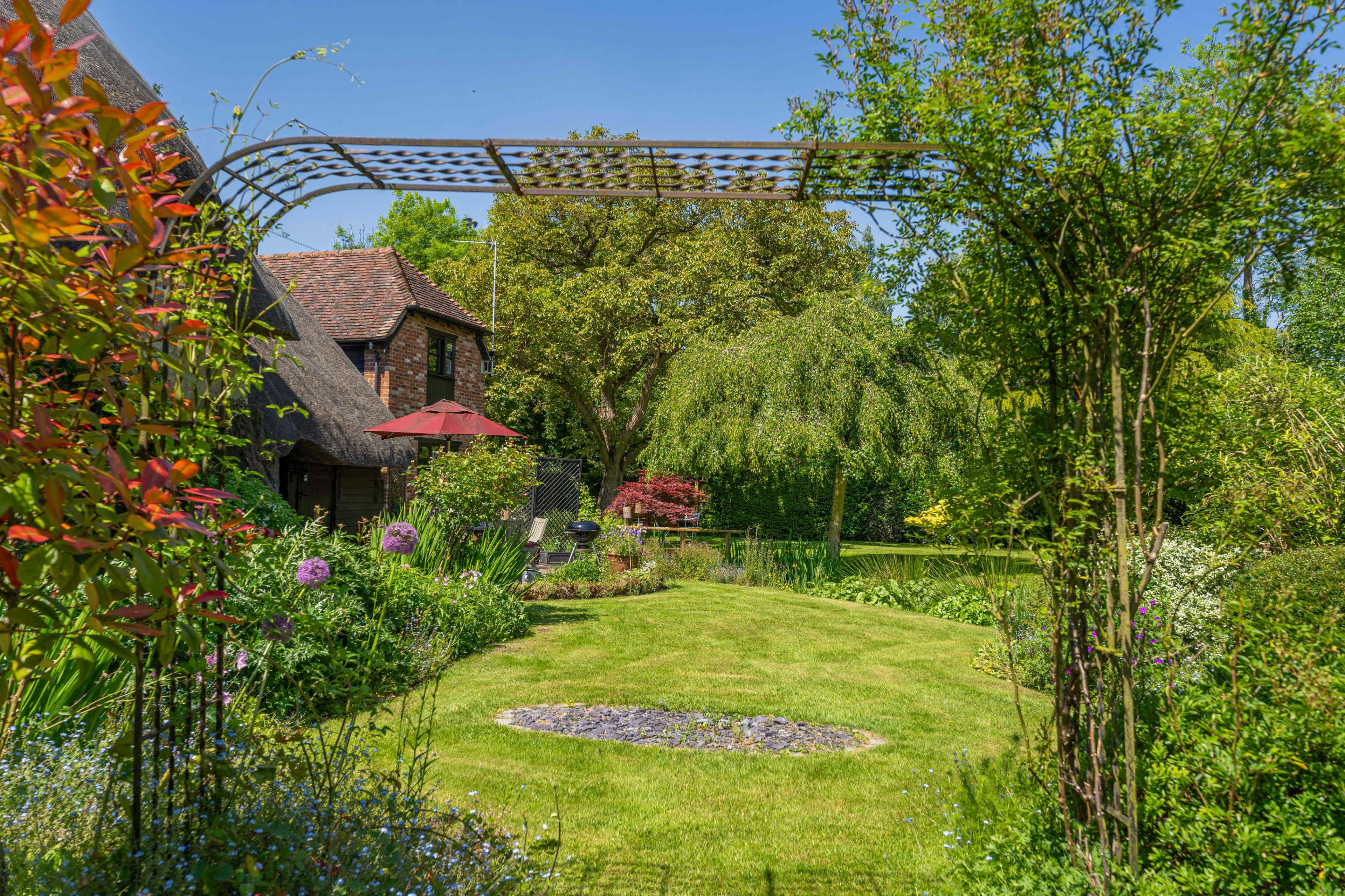 A landscaped garden featuring a metal archway, vibrant flowers, and a thatched house in the background under a clear blue sky.