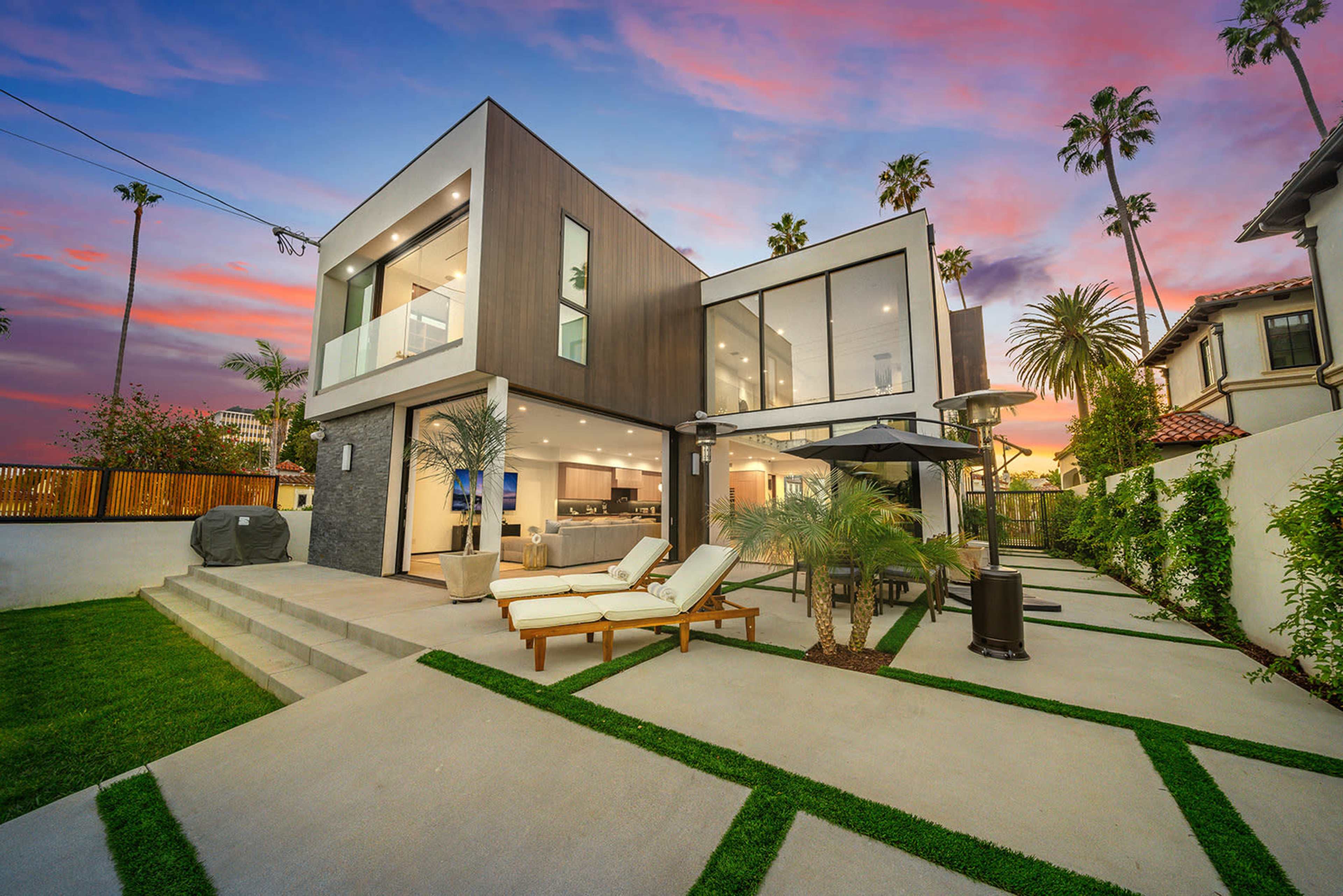 A modern two-story house with large glass windows and a landscaped backyard is set against a colorful sunset sky.