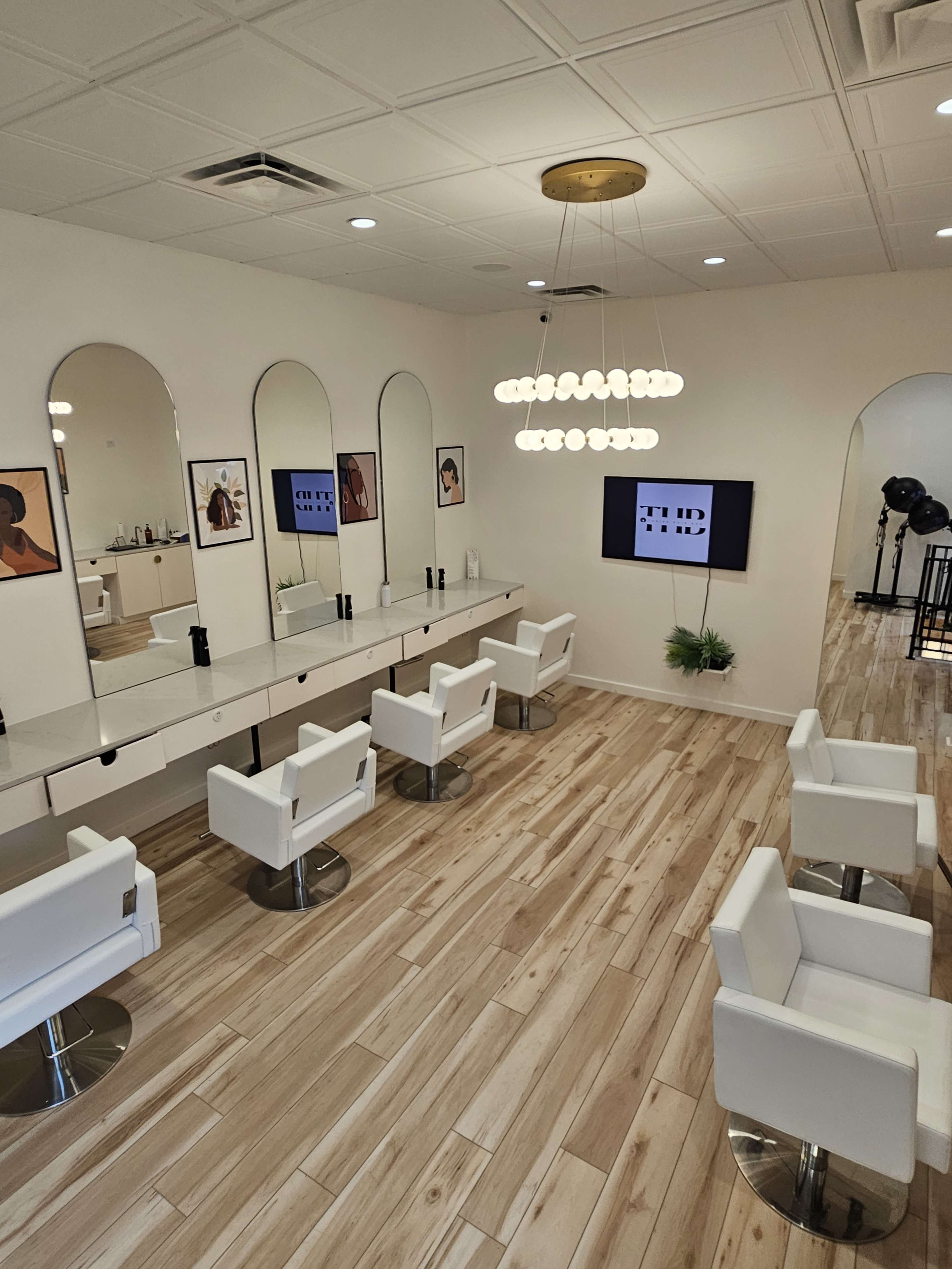 A modern hair salon featuring white chairs, a mirrored wall with framed artwork, and a central light fixture.