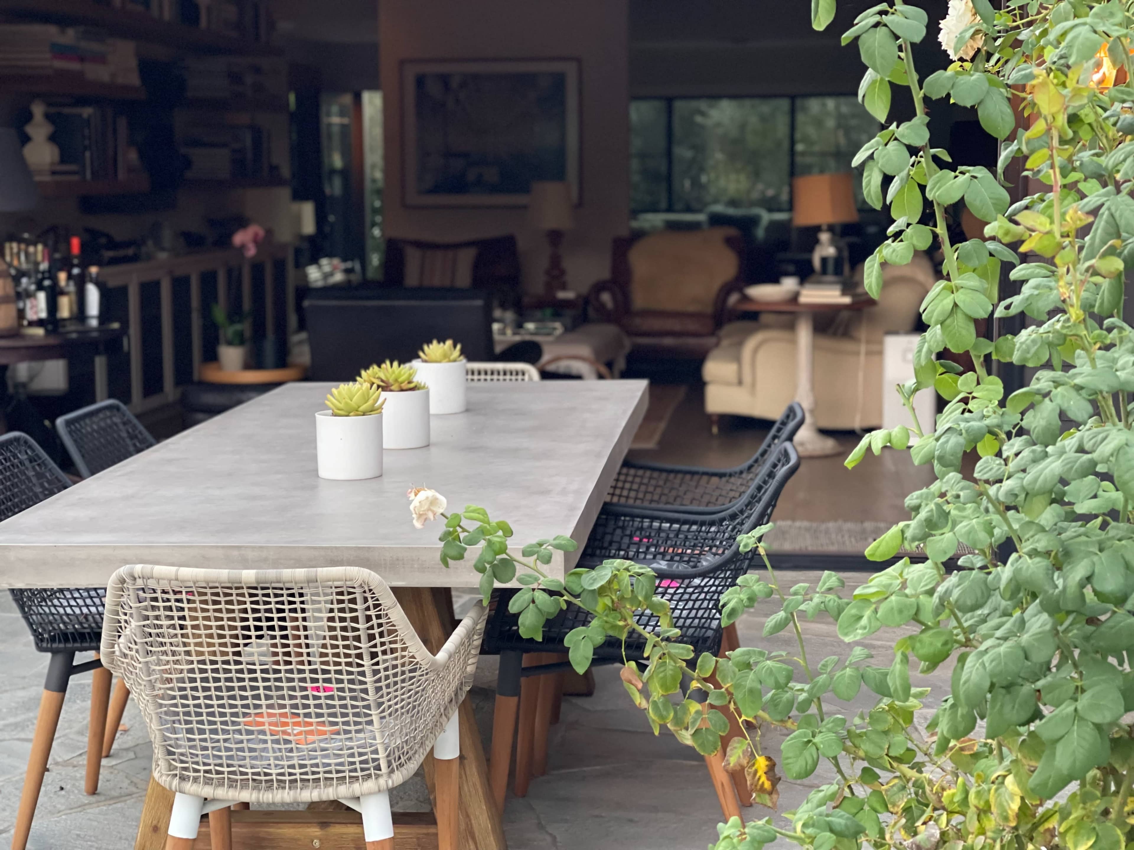 A large stone table is surrounded by chairs, with a view of a living area behind, featuring bookshelves and comfortable seating.