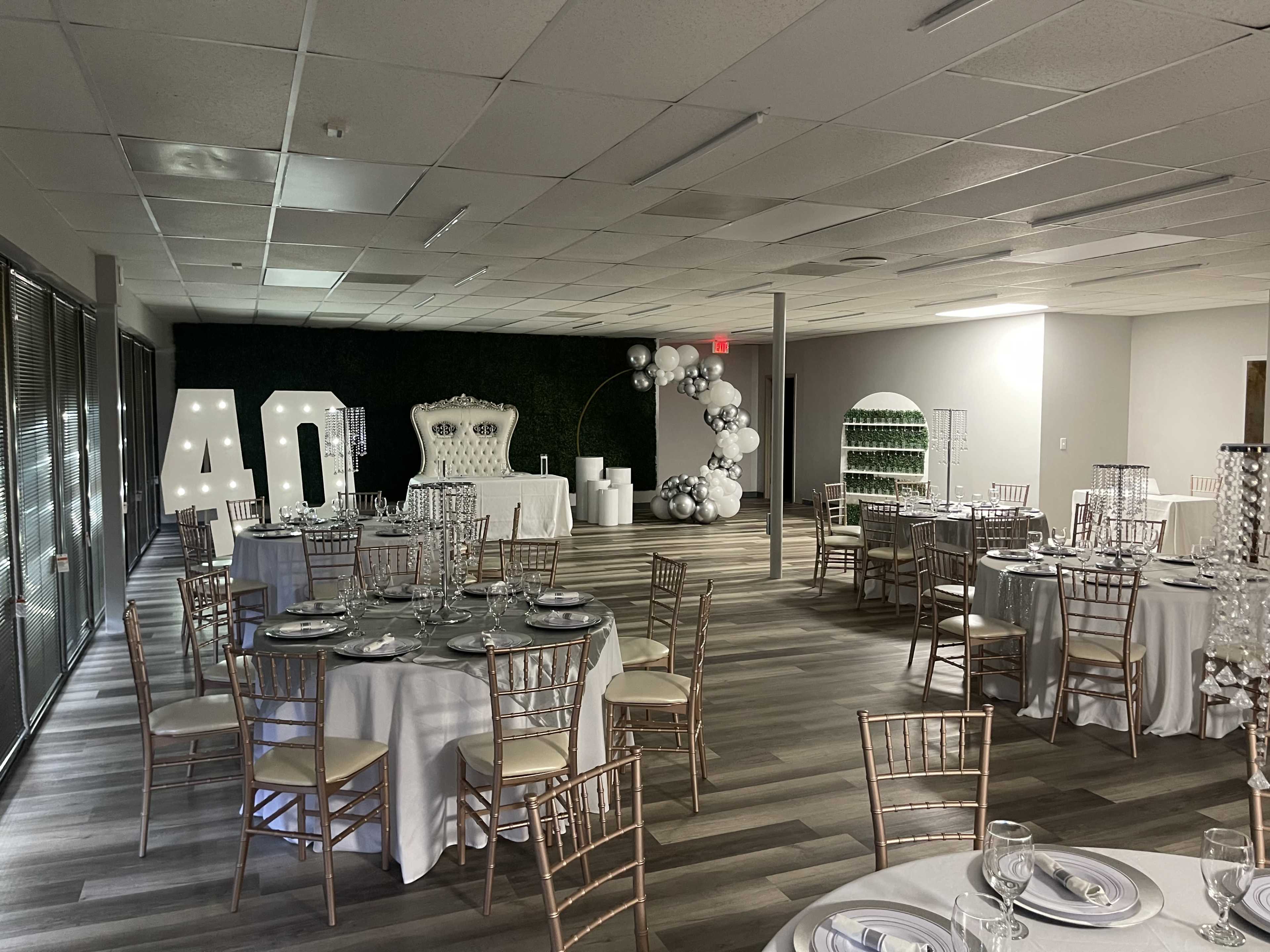 The image shows a banquet hall set up for a 40th birthday celebration, featuring round tables with white tablecloths, gold chiavari chairs, and decorative elements including large number lights and balloon arrangements.