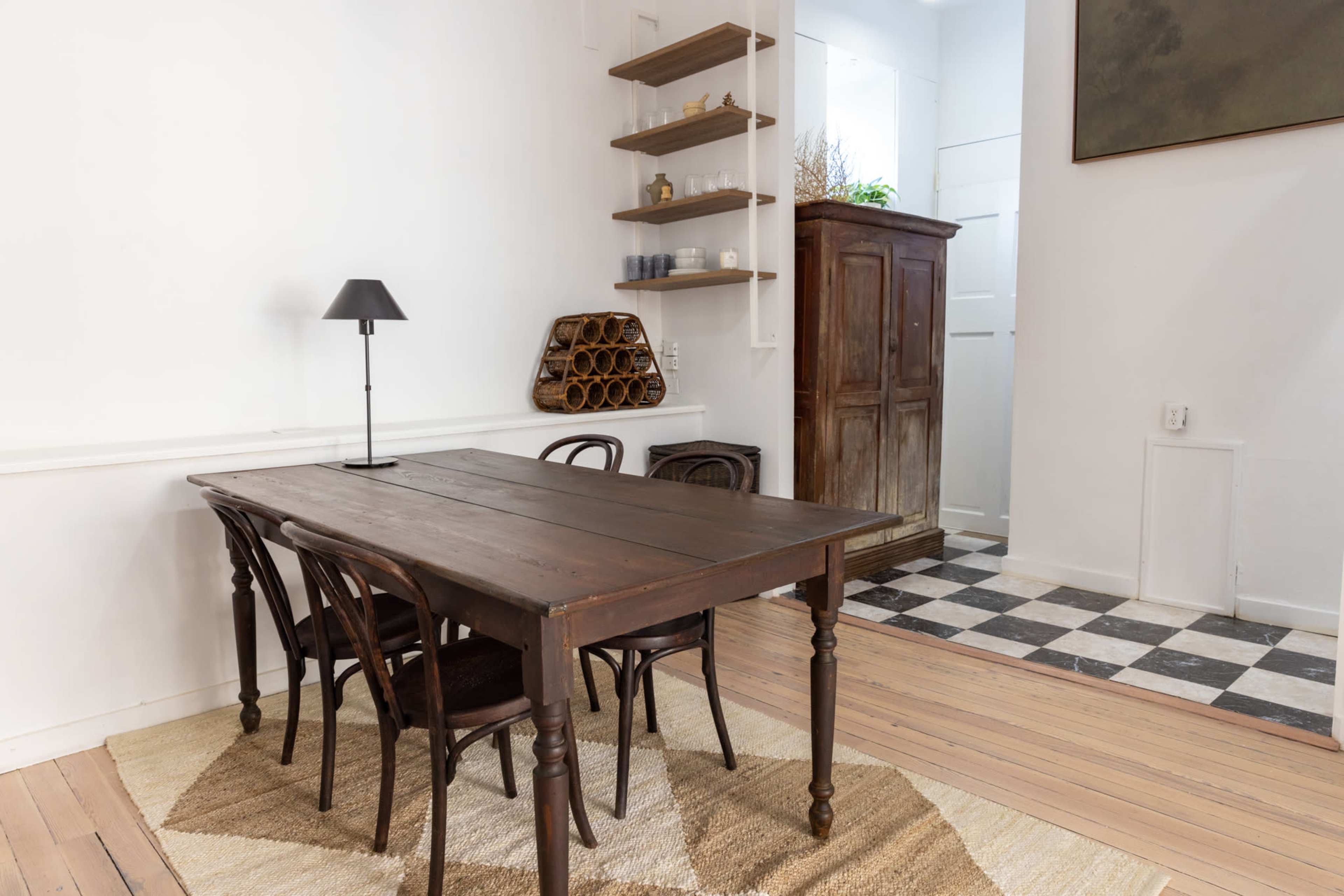A wooden dining table with four chairs sits on a rug in a room with white walls, featuring a small lamp and shelves on one side, and a patterned floor on the other.