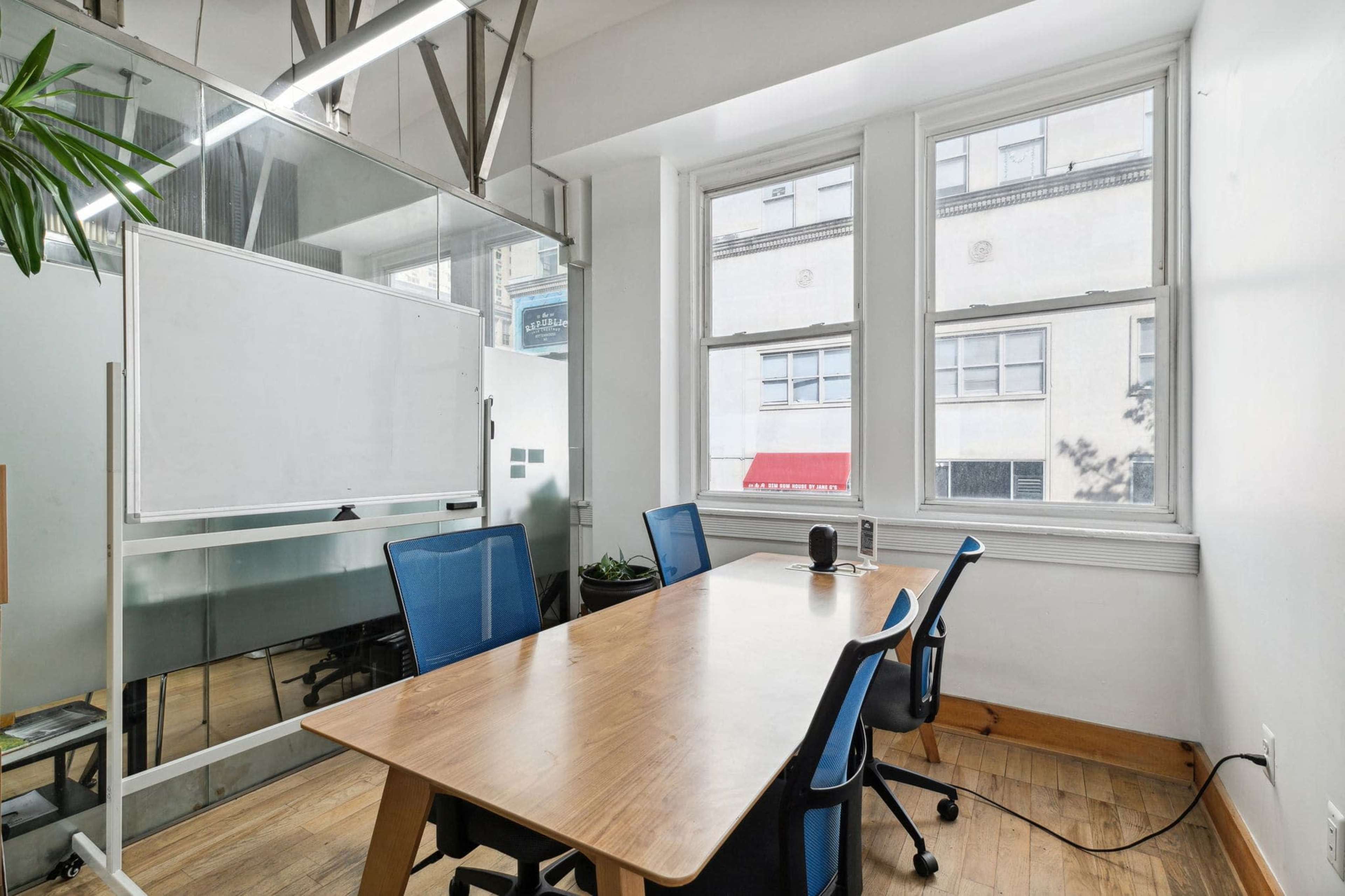 A conference room features a wooden table with three blue chairs, a whiteboard on wheels, and large windows allowing natural light in.
