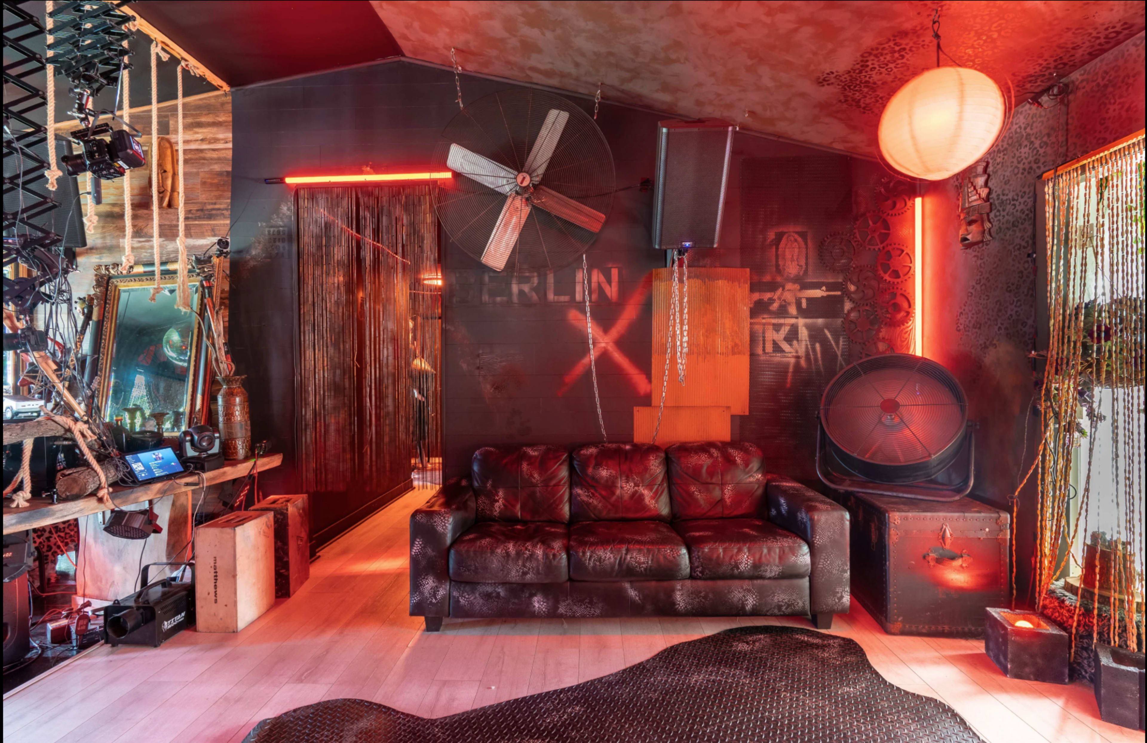 The image depicts an eclectic room featuring a dark leather sofa, industrial decor, various lighting elements, and an intricately designed wall.