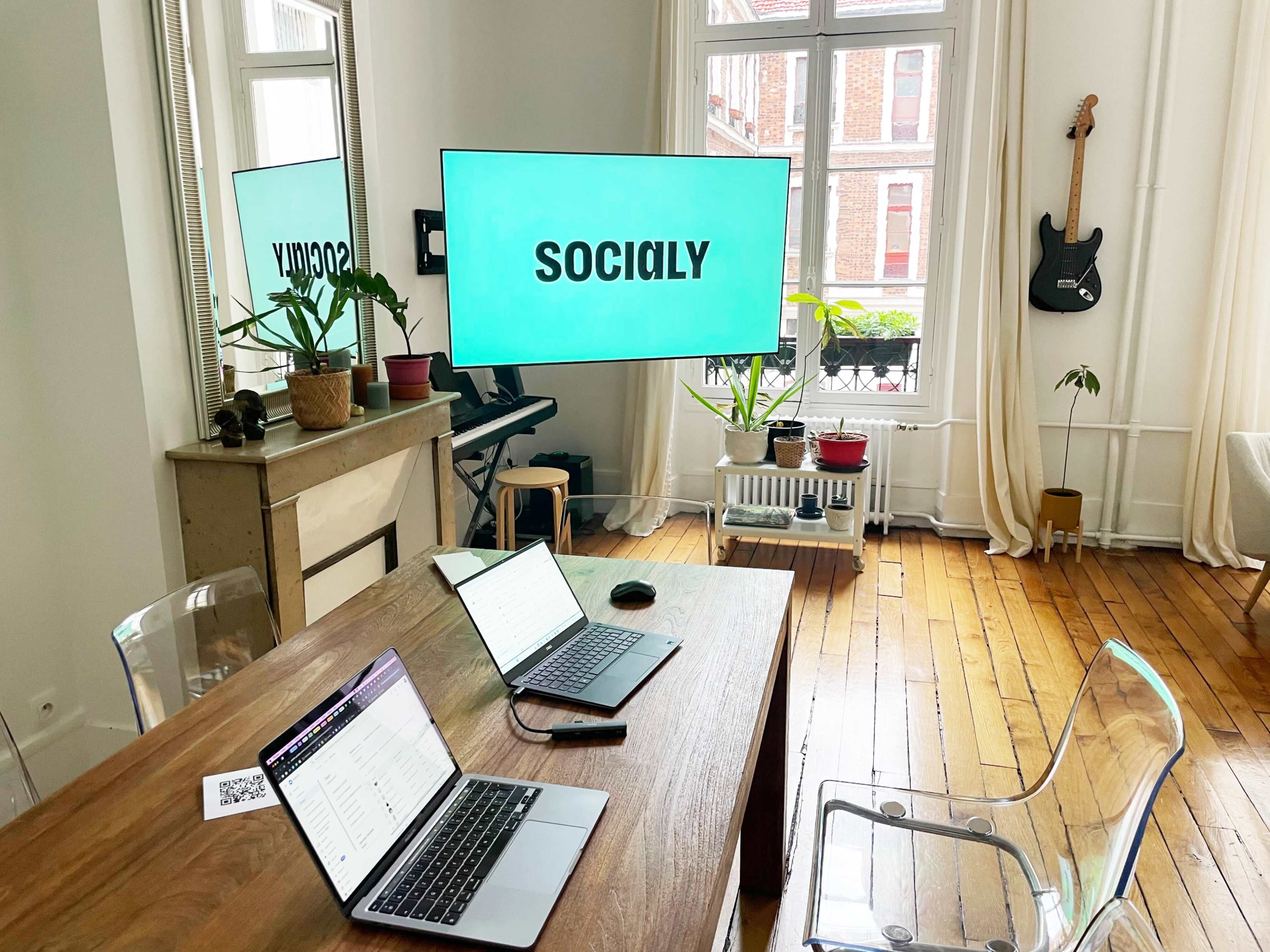 A modern workspace features two laptops on a wooden table, a large TV displaying the word "SOCIALLY," plants, and a guitar on the wall.