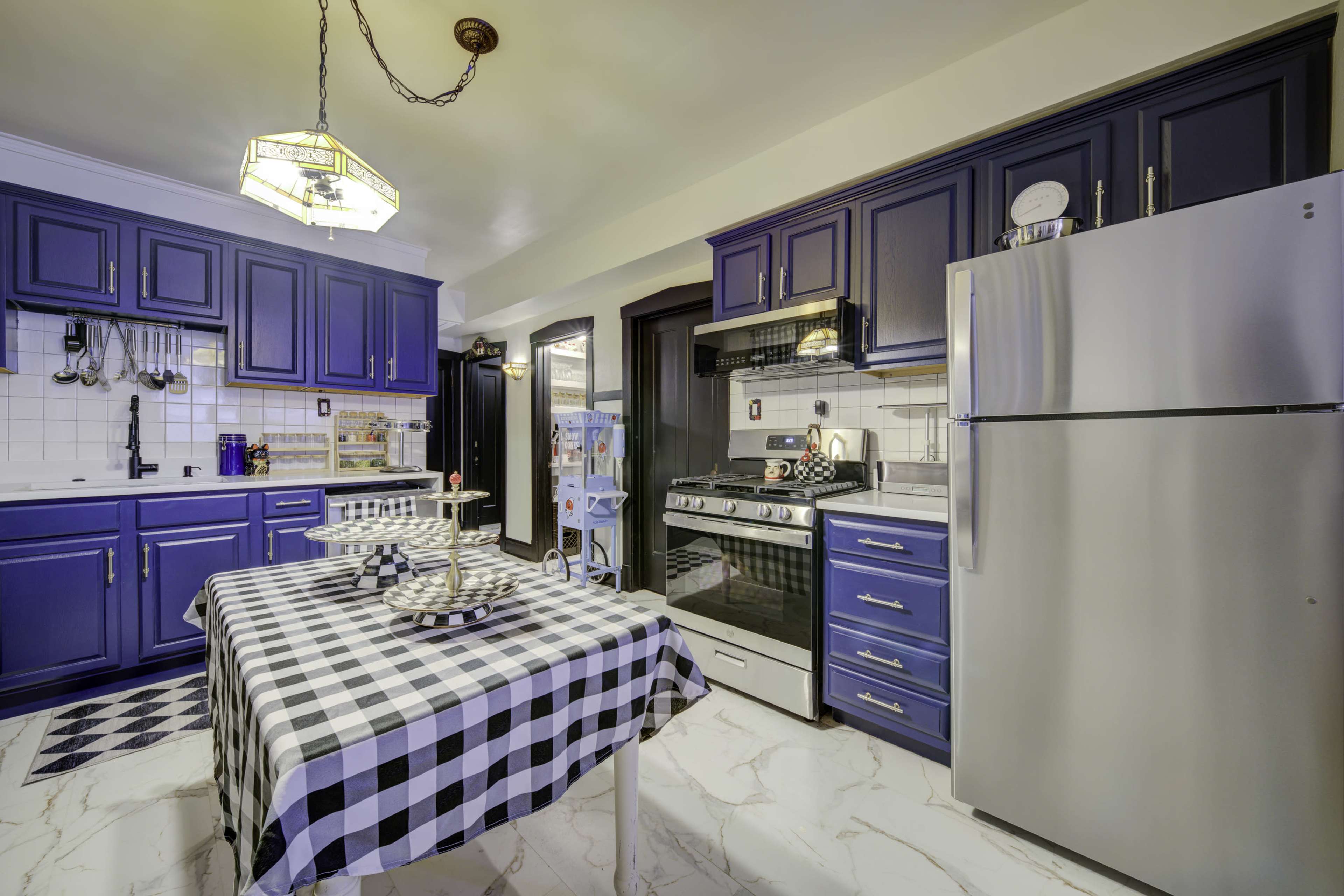 A modern kitchen features blue cabinets, stainless steel appliances, and a checkered tablecloth on a dining table.
