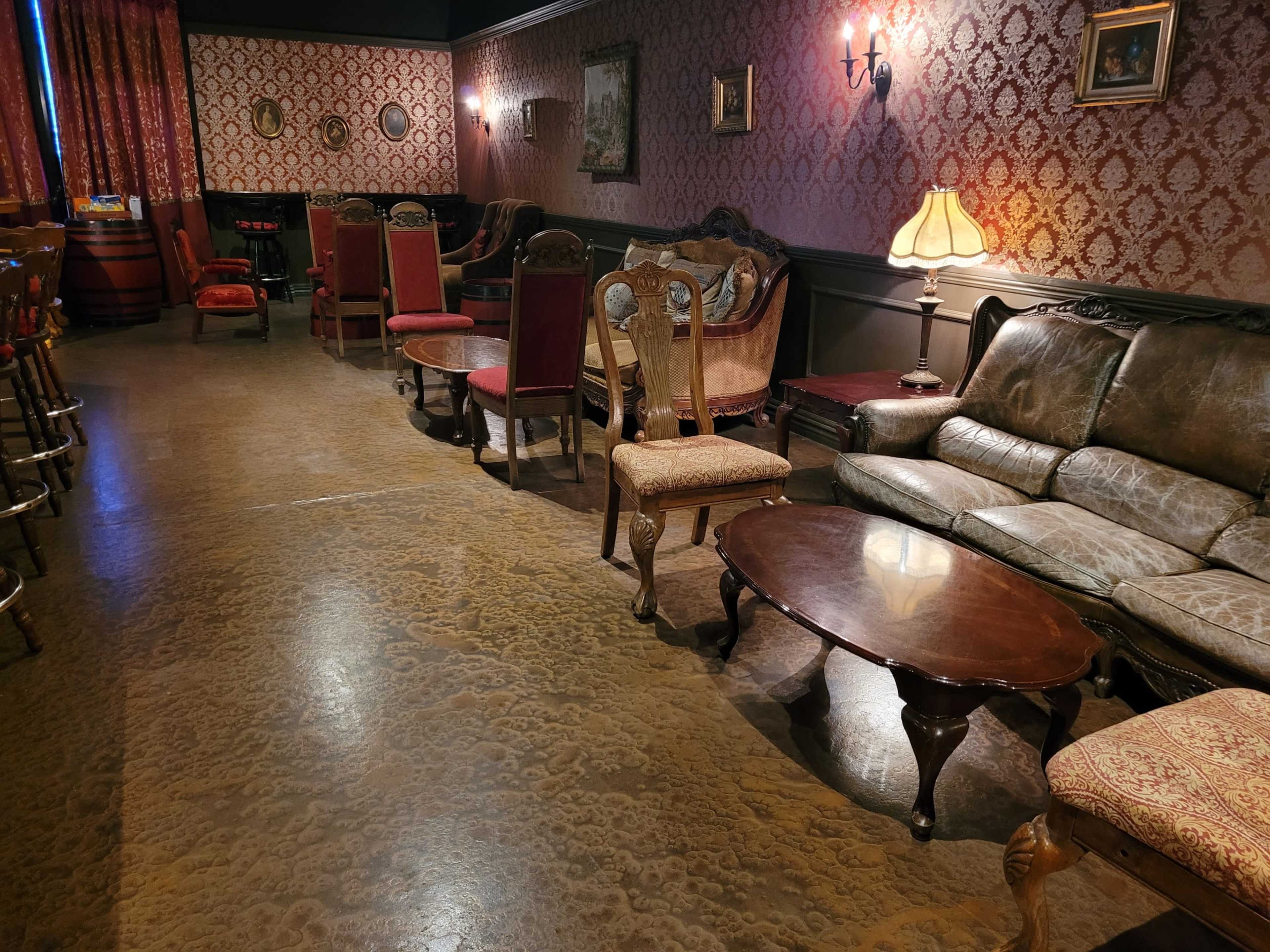 A vintage-style lounge area with ornate furniture, including a leather sofa, wooden chairs, and a small round table, surrounded by patterned wallpaper and soft lighting.