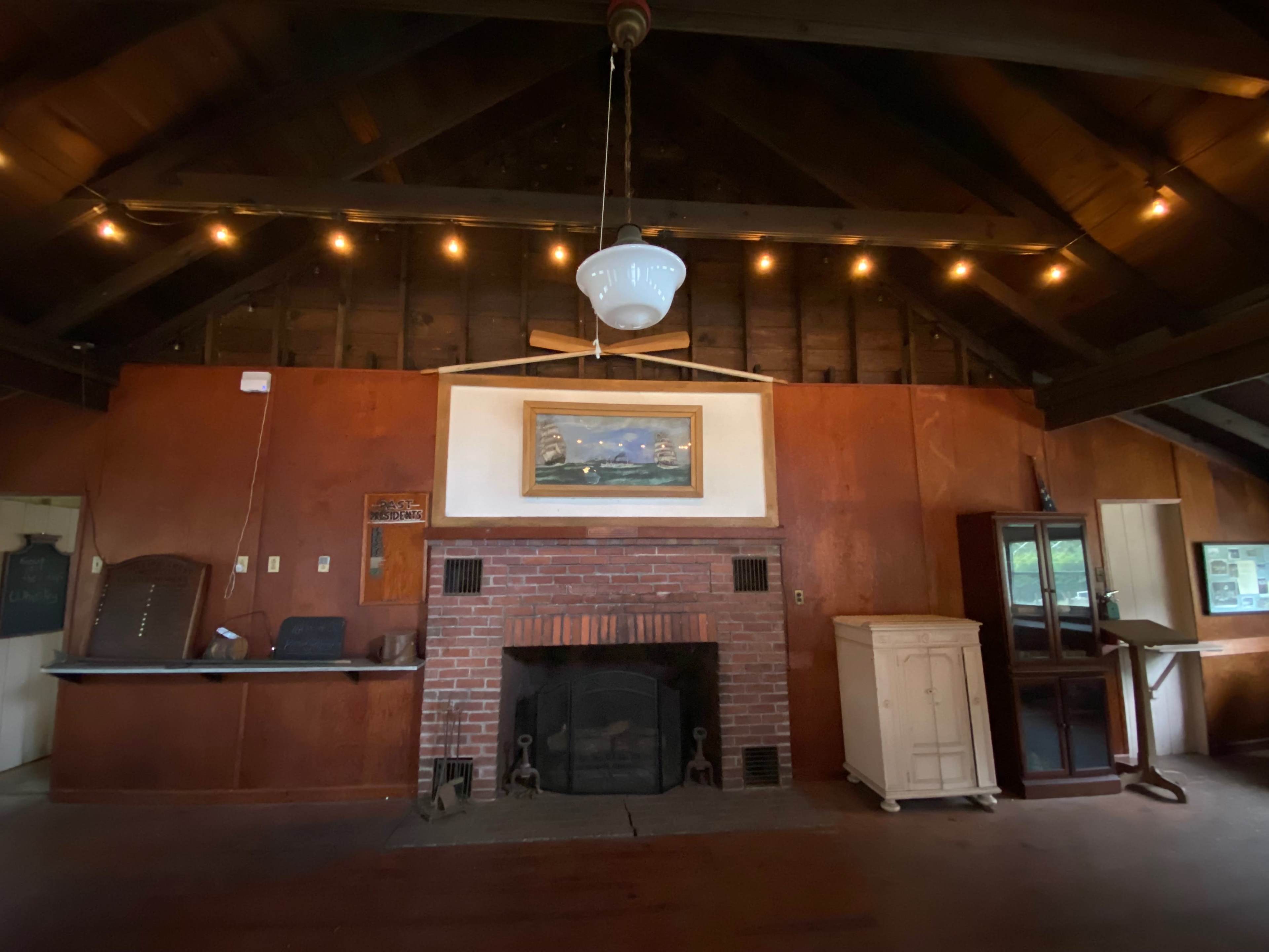 The interior of a rustic room features a brick fireplace, wooden beams, and a painting above the mantle.