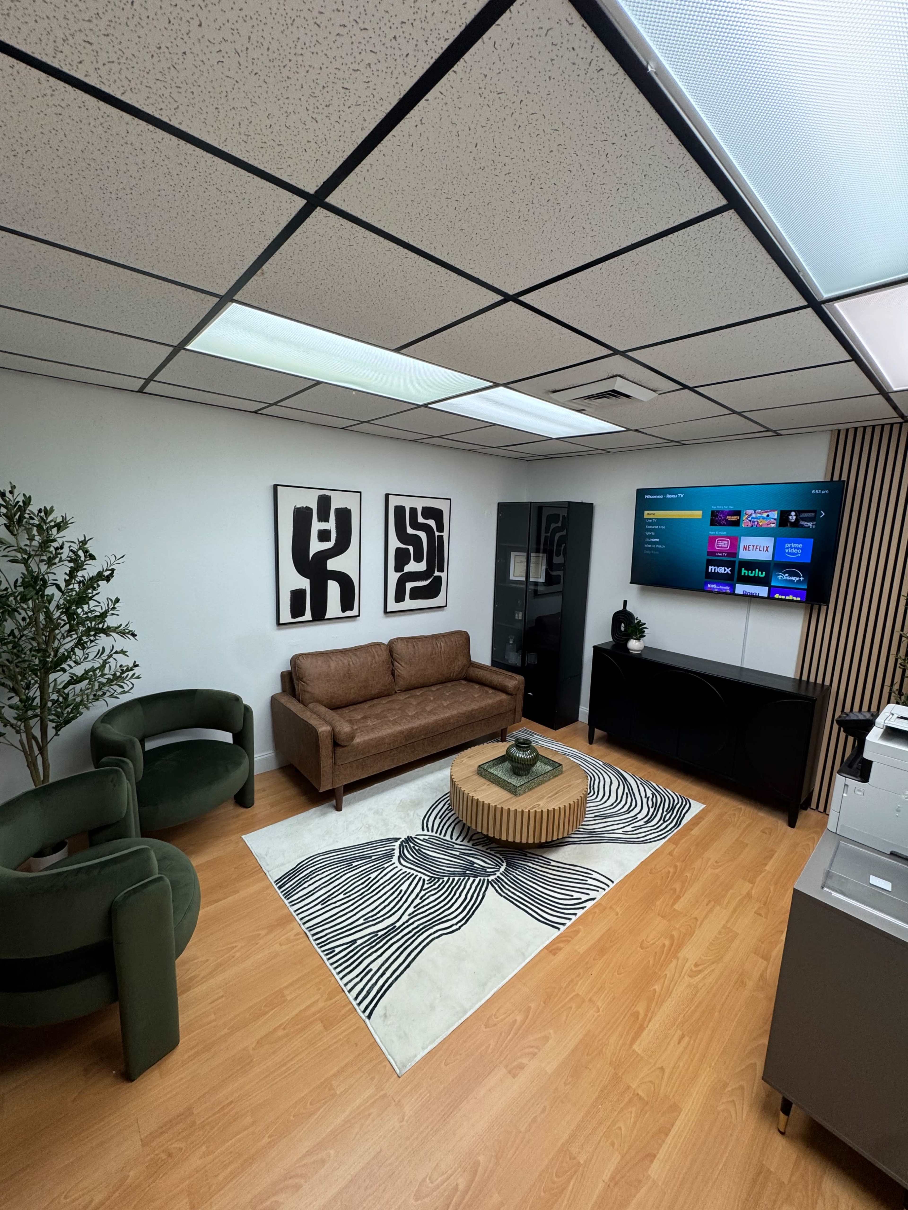 The image shows a modern office seating area with a brown sofa, two green chairs, a round coffee table, a patterned rug, a television on the wall, and a small indoor plant.