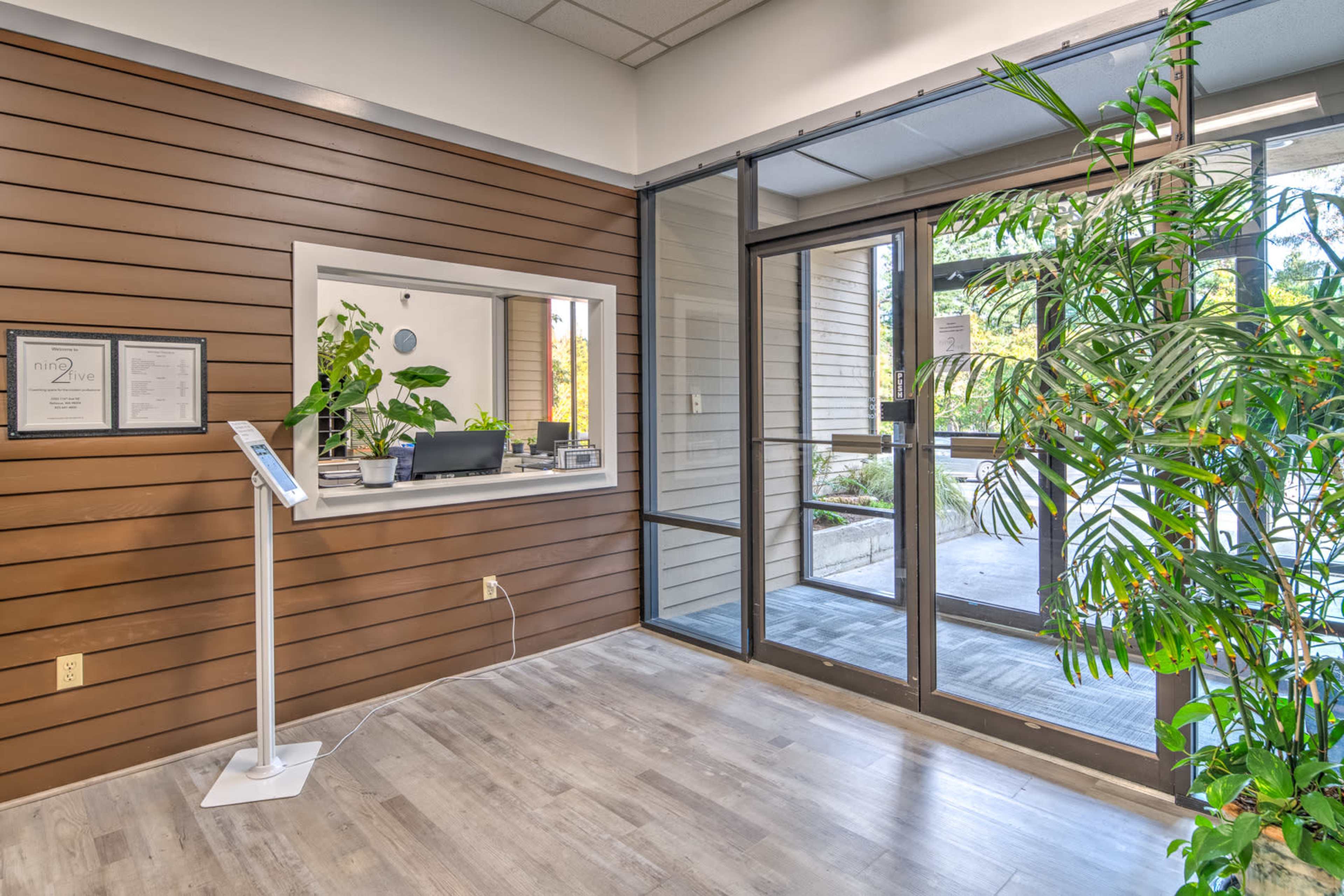 A reception area with a glass door, a desk with plants, and a digital information stand.