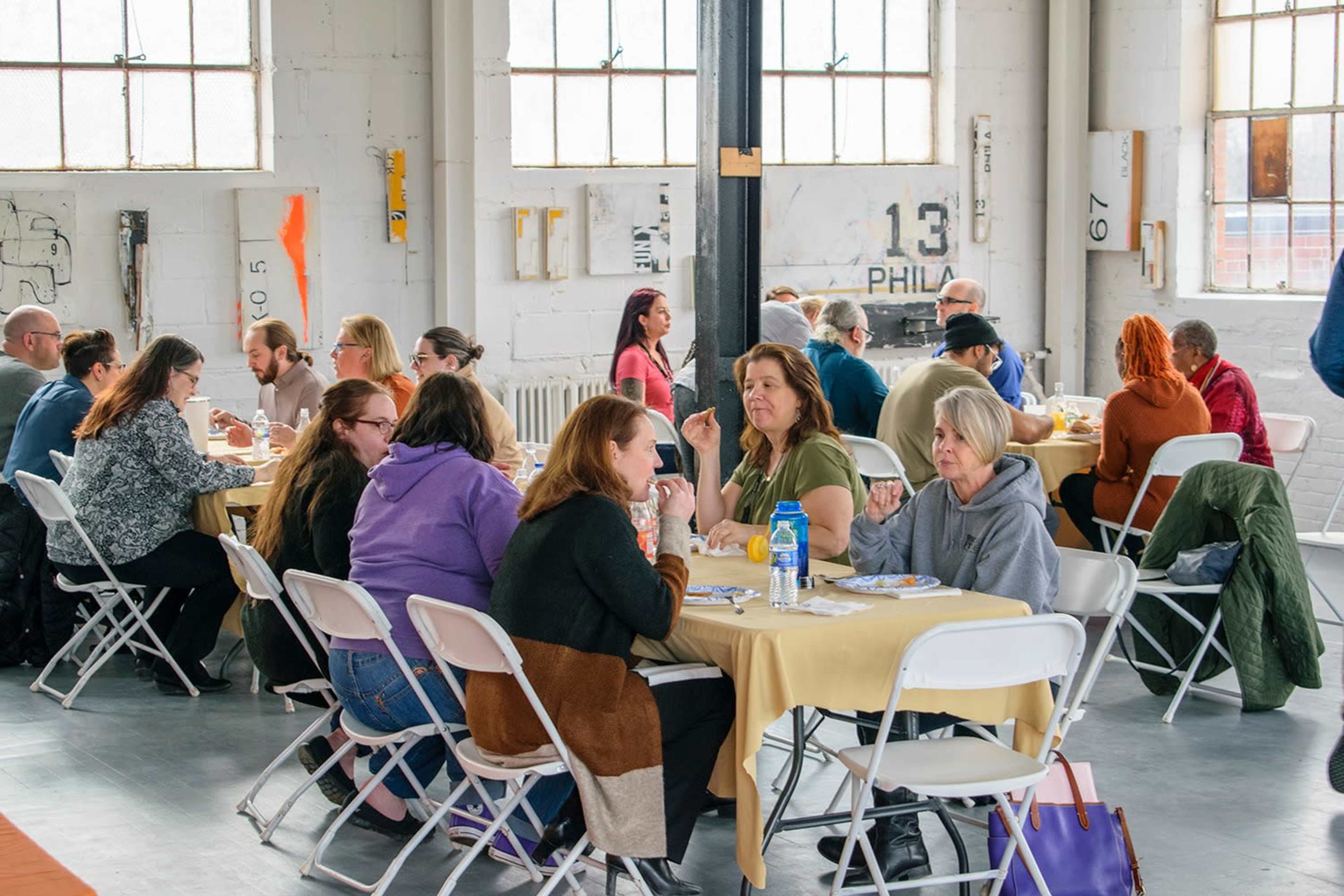 A group of people is seated at tables, eating and conversing in a spacious, industrial-style room with large windows and minimal decor.