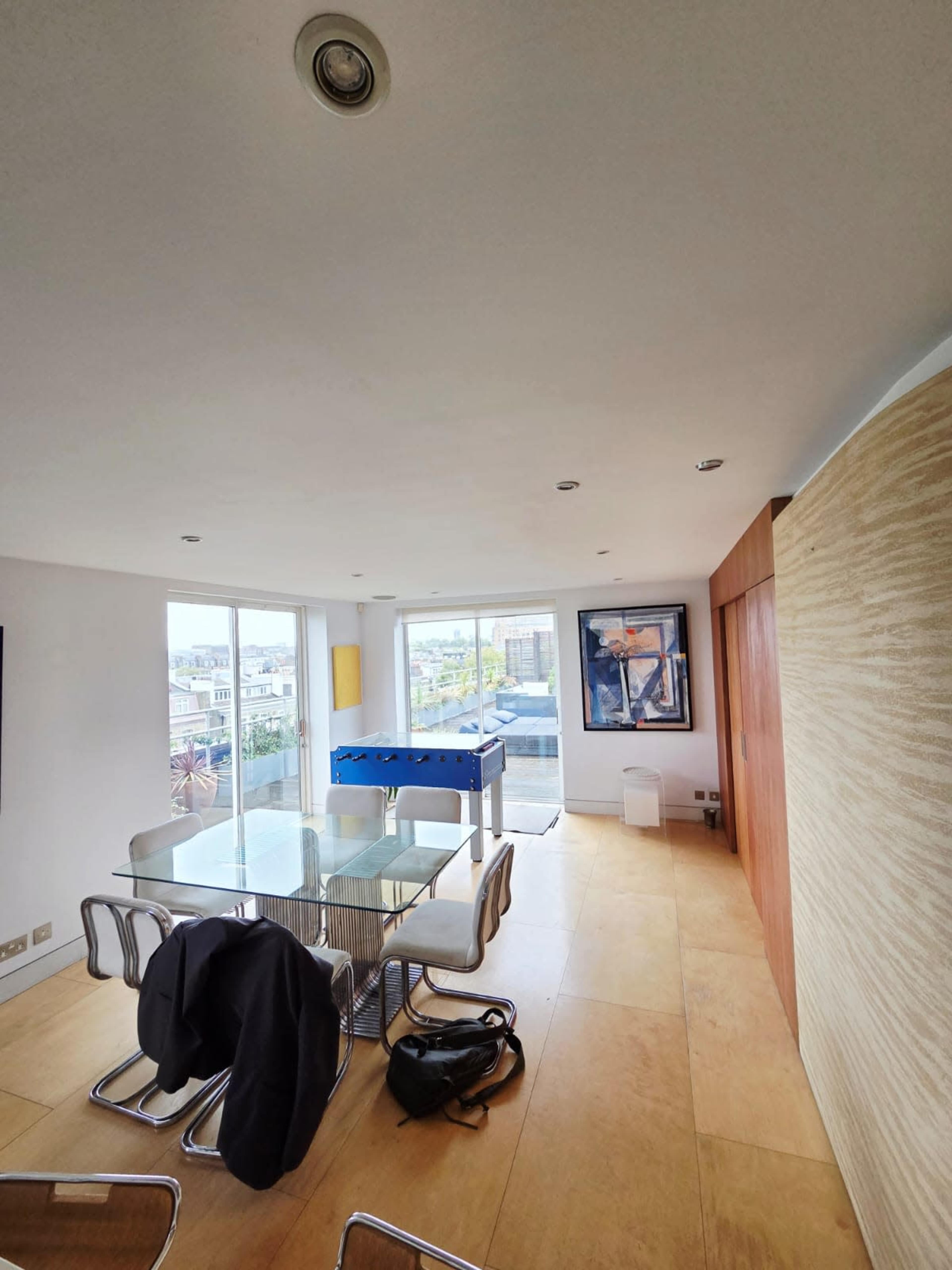 The image shows a modern dining area with a glass table and chairs, large windows providing natural light, and a visible outdoor view.