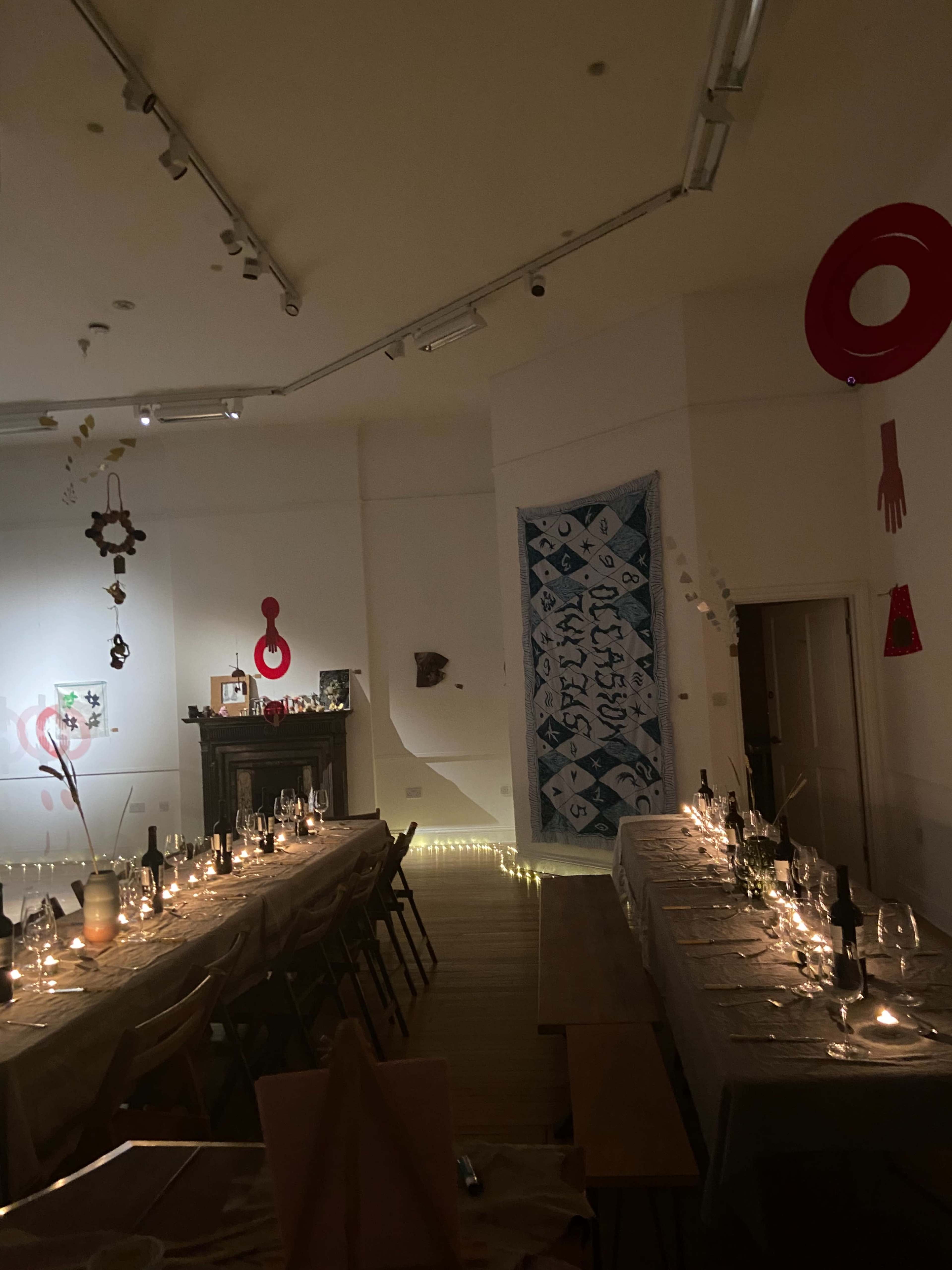 A long dining table is set with candles and wine bottles under dim lighting, surrounded by decorative art pieces on the walls.