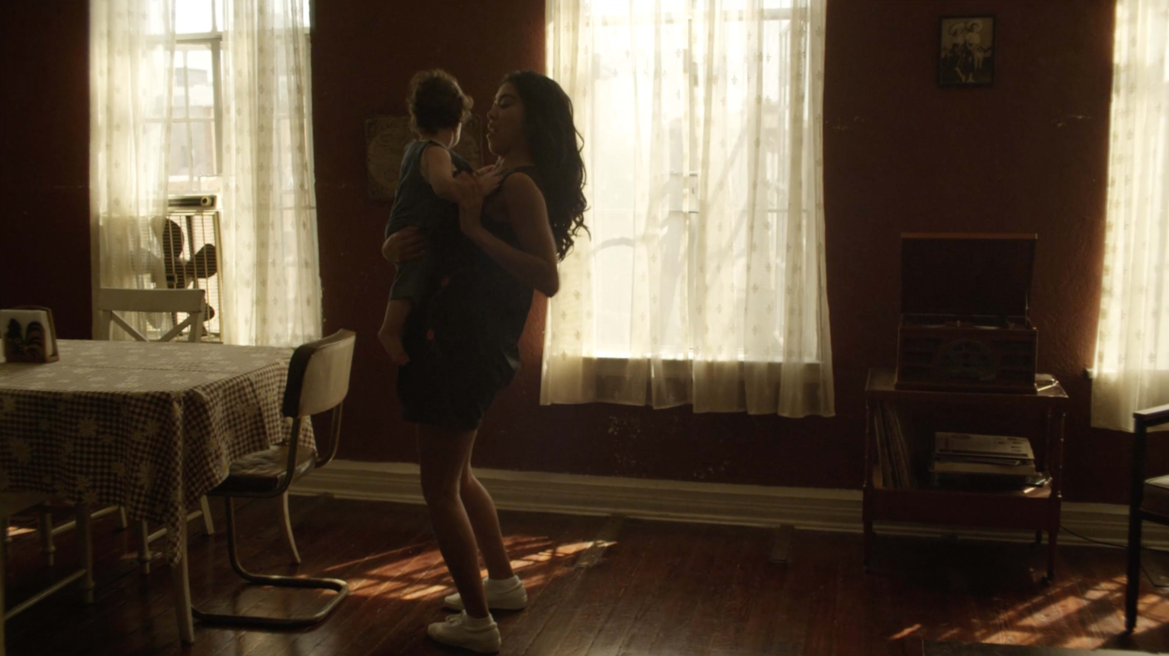 A woman holds a child while standing in a sunlit room with a table and a record player.