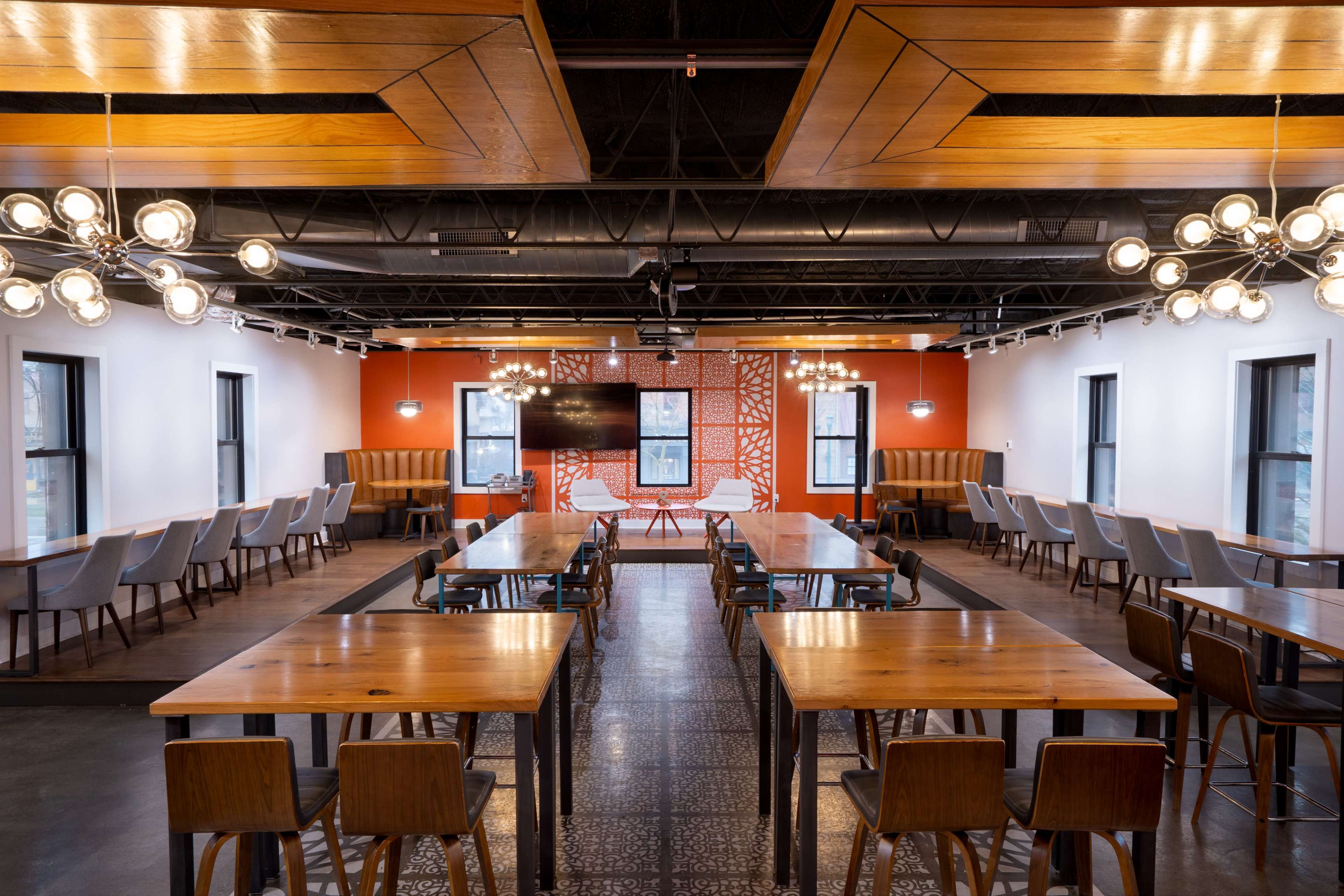 The image shows a spacious, modern meeting room with rows of tables and chairs, large windows, and a wall featuring an orange design.