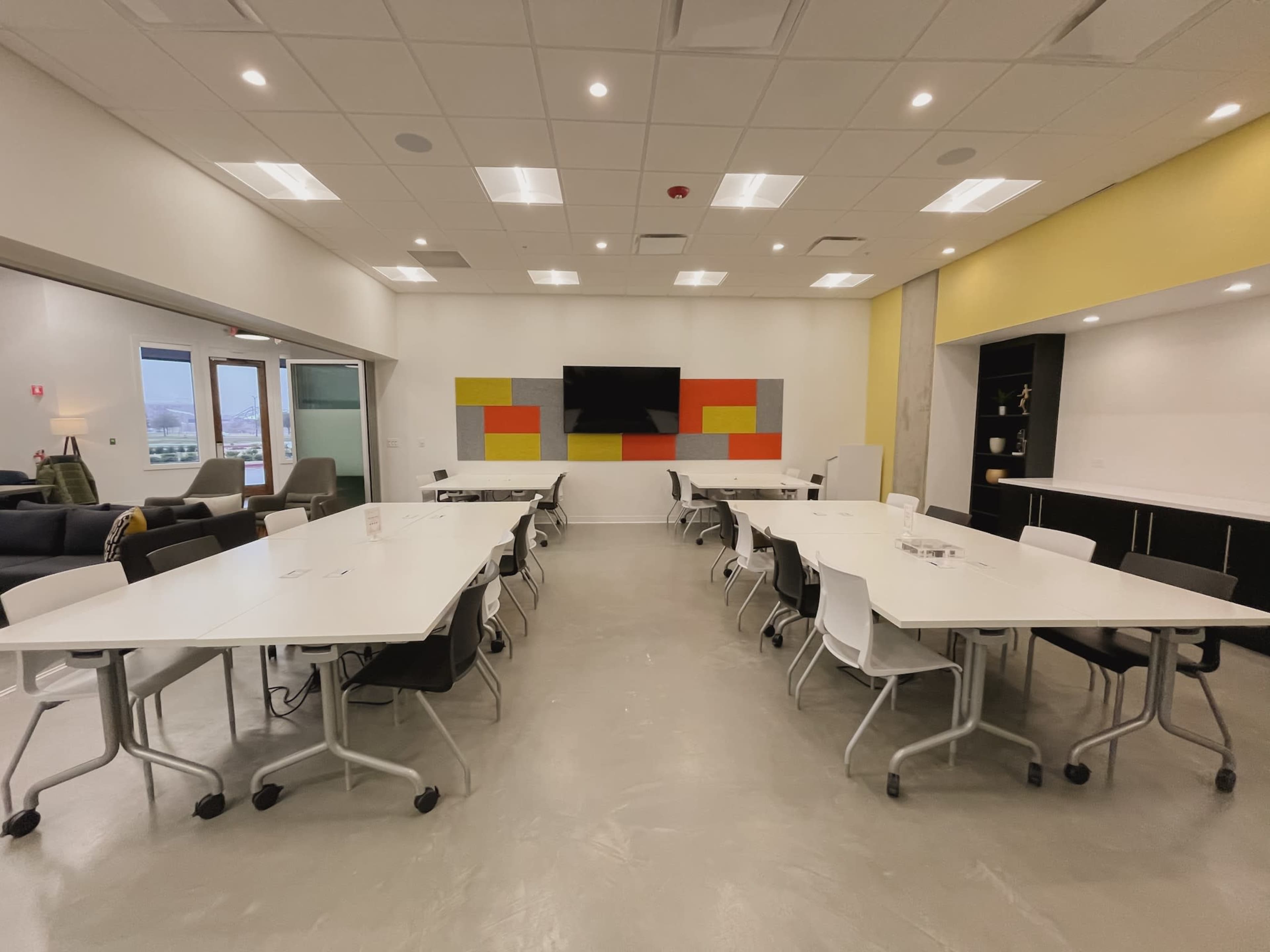 A spacious meeting room features multiple white tables arranged in a U-shape, with a wall-mounted screen and colorful acoustic panels in the background.