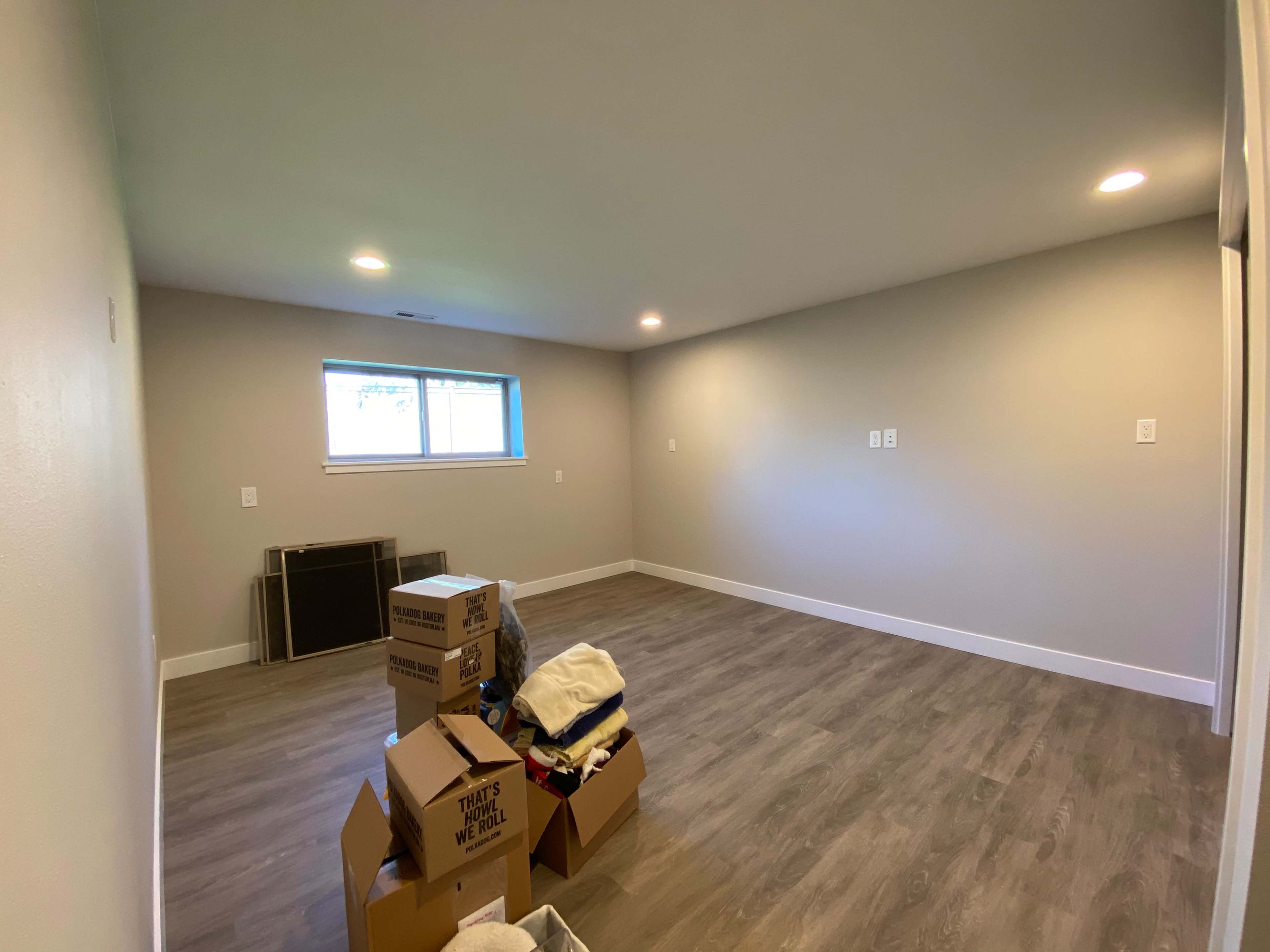 A spacious, empty room with pale gray walls, light brown flooring, and a stack of boxes and clothing in one corner.