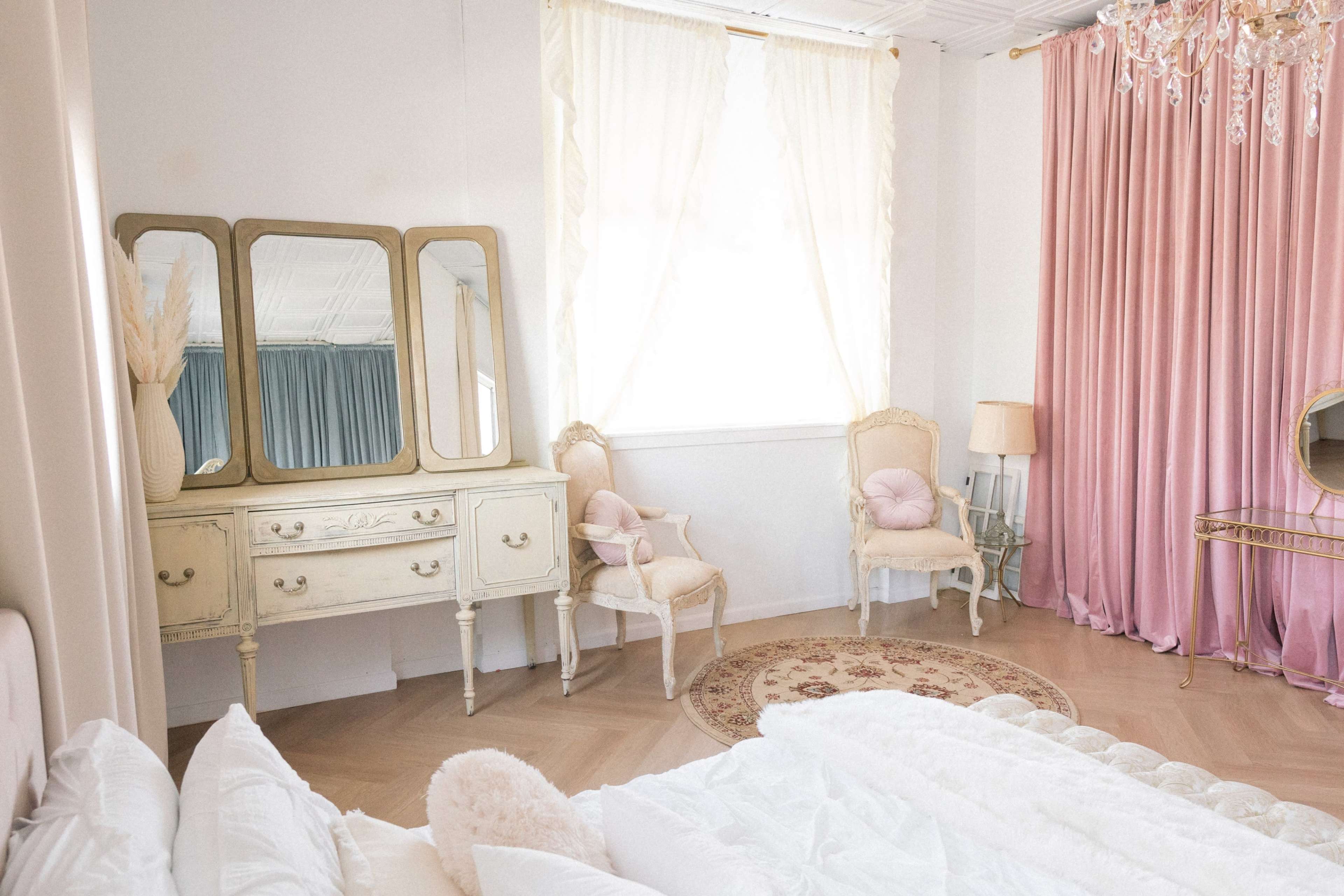 A bright bedroom features a bed with white bedding, an ornate dresser with three mirrors, two armchairs, a decorative rug, and large pink curtains.